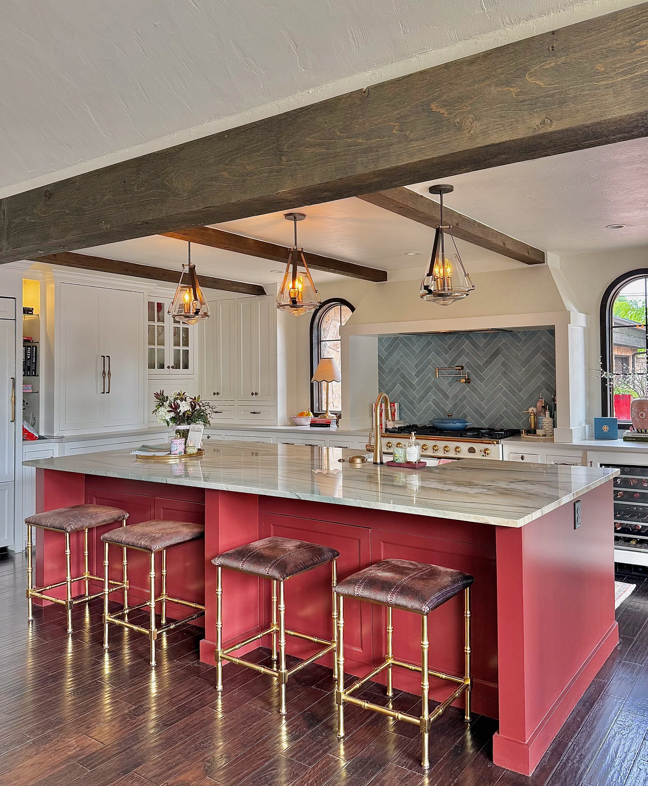 Modern kitchen with white cabinets, a marble island with a pink base, gold and burgundy bar stools, dark hardwood floors, exposed ceiling beams, and pendant lights.