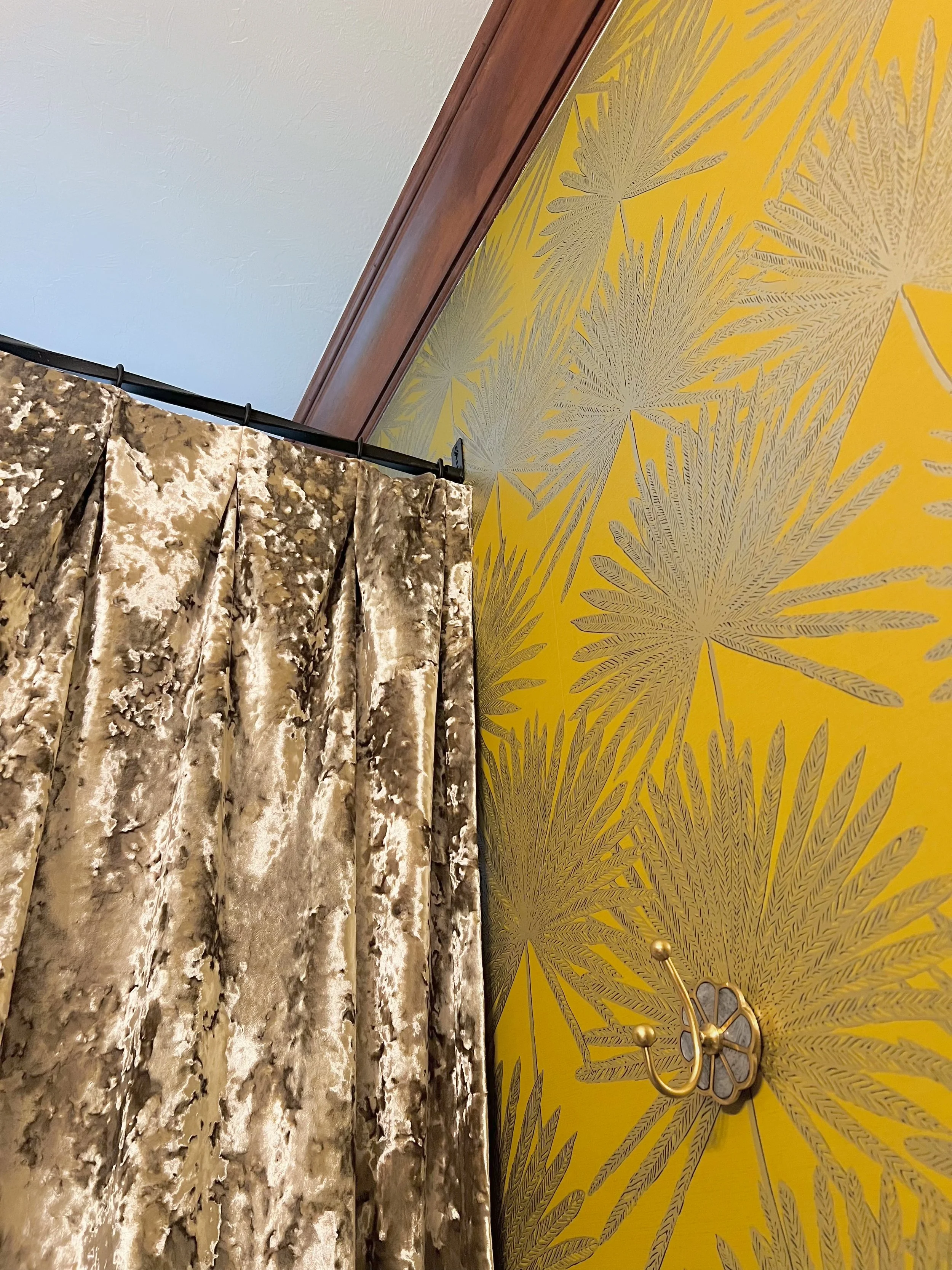 A part of a room corner with a yellow wall featuring a tropical leaf pattern in gold, a velvet curtain with a marble-like beige and brown pattern, and a decorative gold wall hook.