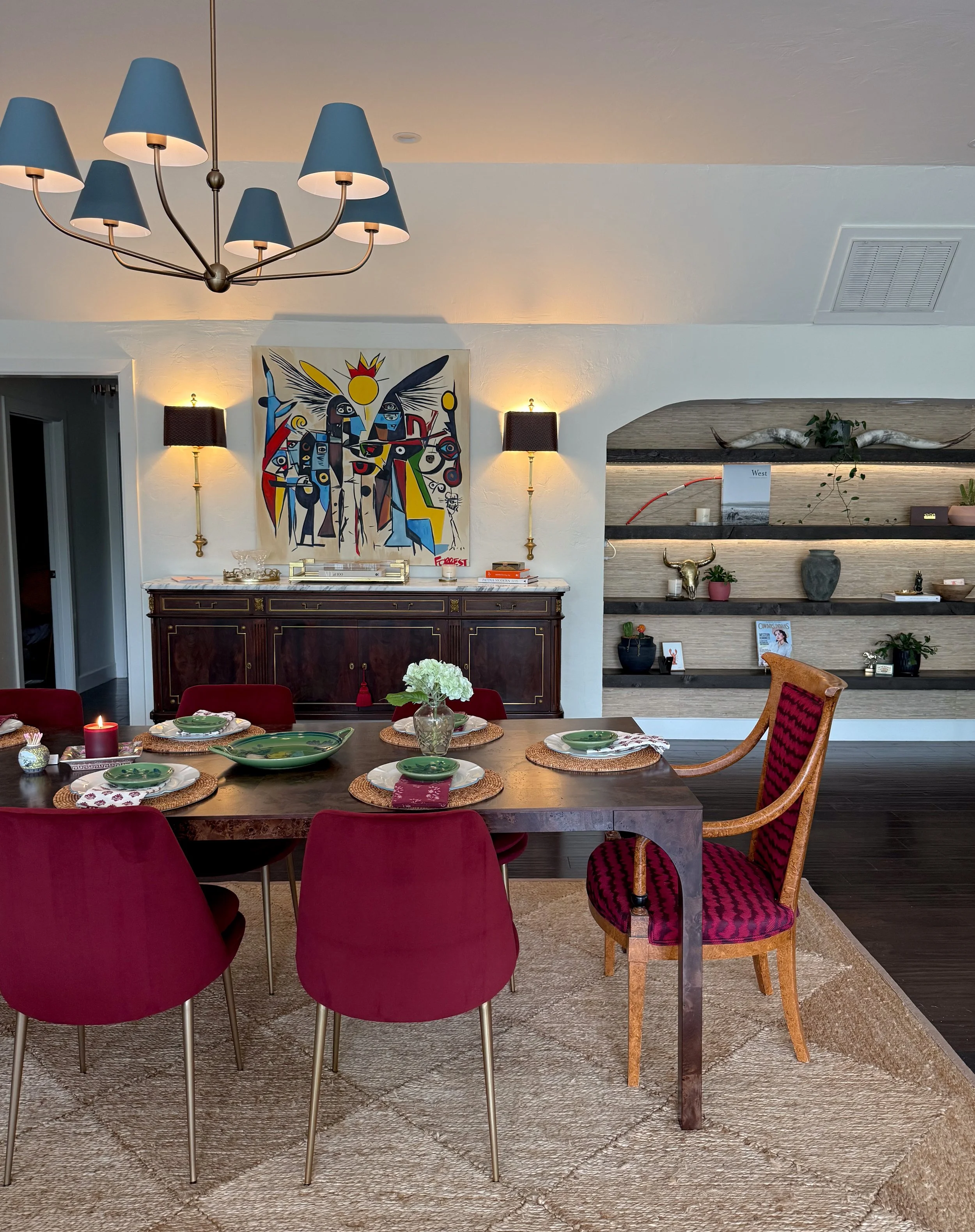 A dining room with a wooden table set with green plates, white napkins, and a vase of white flowers. Red velvet chairs surround the table, with a unique wooden armchair at the end. A large abstract painting hangs above a dark wooden sideboard. Two wa