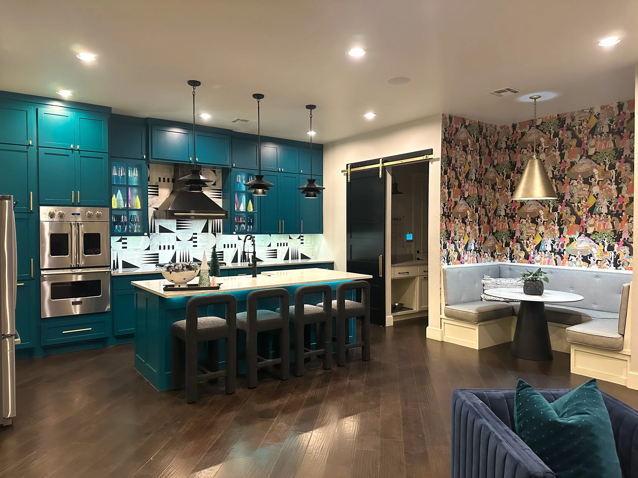 Modern kitchen with teal cabinets, black pendant lights, a patterned backsplash, stainless steel appliances, a central island with barstools, and a cozy corner nook with a rounded bench and a small round table, decorated for the holidays.