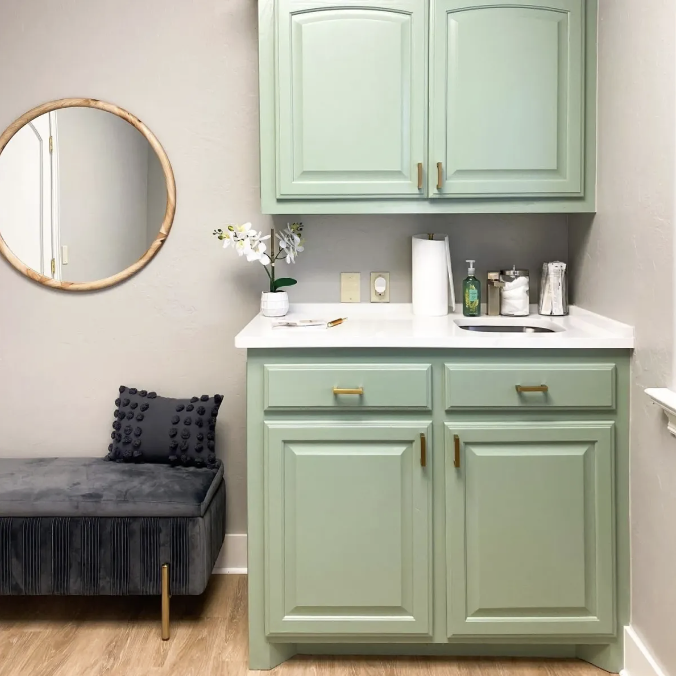 A small room with a mint green cabinet and upper cabinets, a white countertop with a potted orchid and various small items, a gray upholstered bench with a decorative pillow, a round mirror on the wall, and wood flooring.
