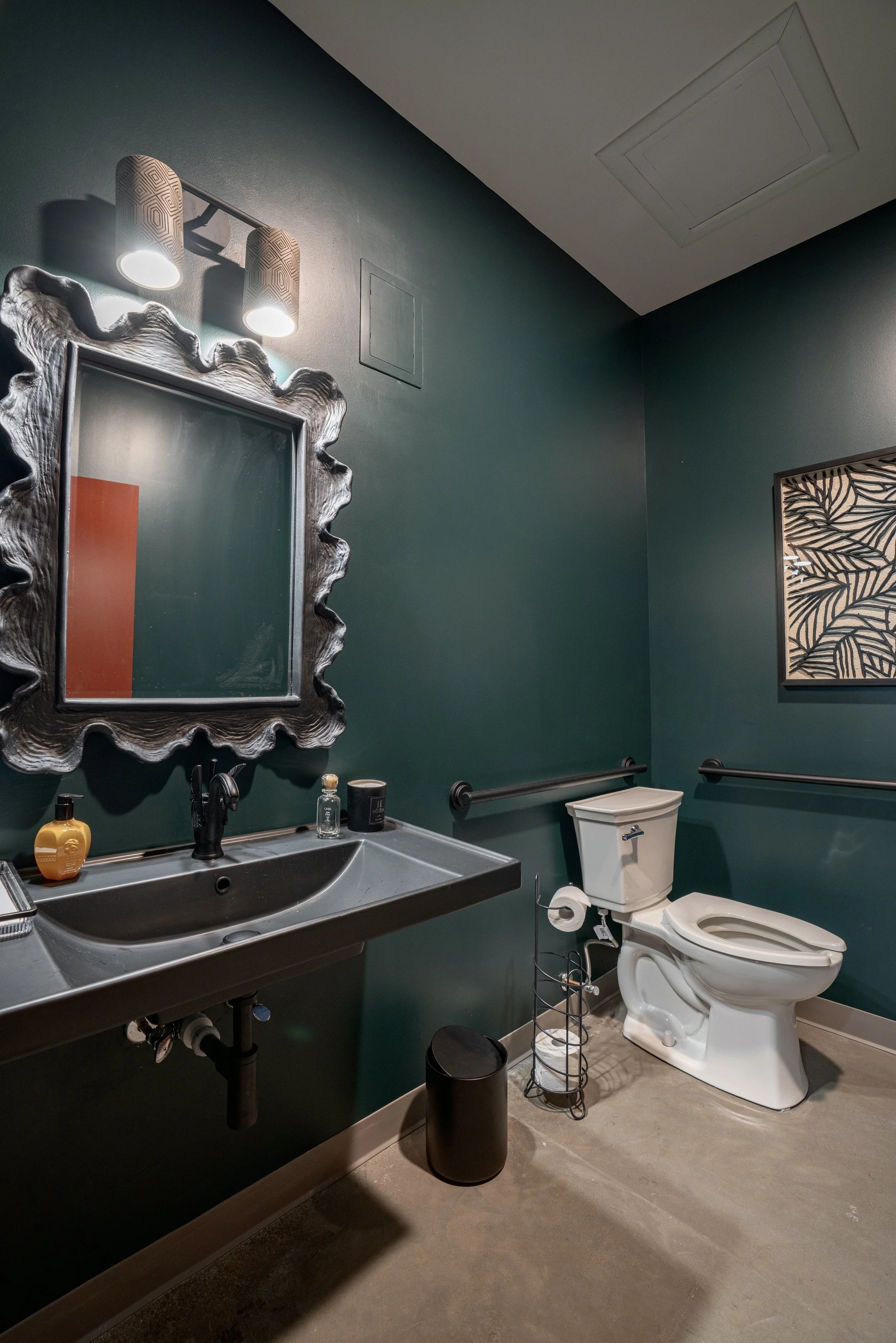 A bathroom with a dark green wall, a black sink with a mirror above, a toilet with a toilet paper holder, wall art with leaves, and a small trash can.