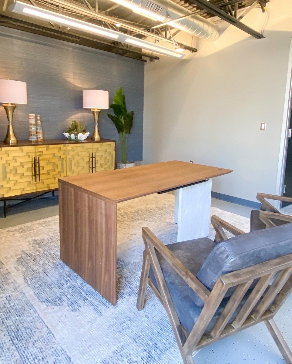Modern office conference room with a wooden desk, four chairs, a yellow sideboard, two table lamps, and a tall green plant.