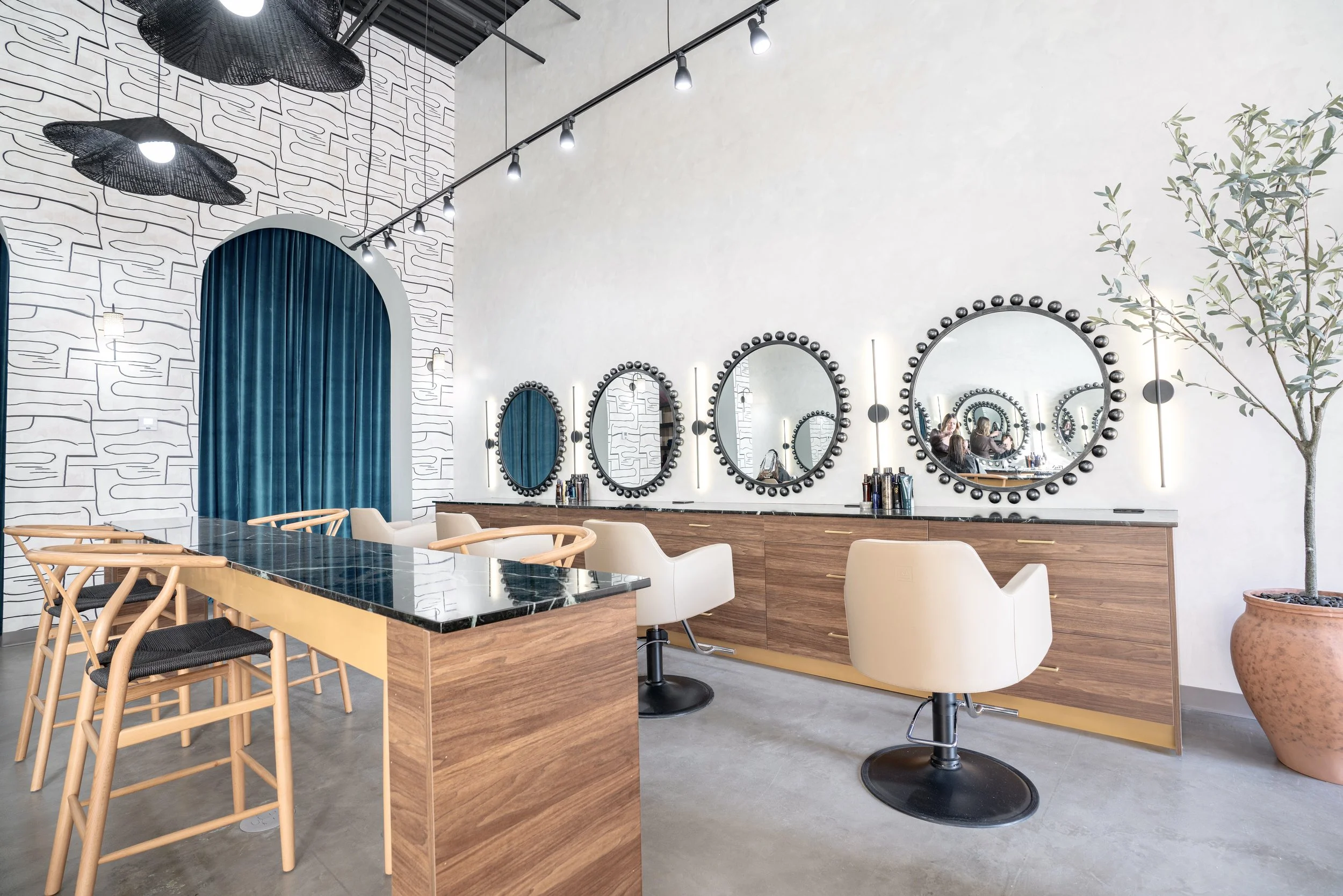 Modern hair salon interior with a row of large mirrors with black circular frames, white salon chairs, a black marble countertop, decorative lighting, a potted plant, and a seating area with high wooden chairs and a marble-topped table.
