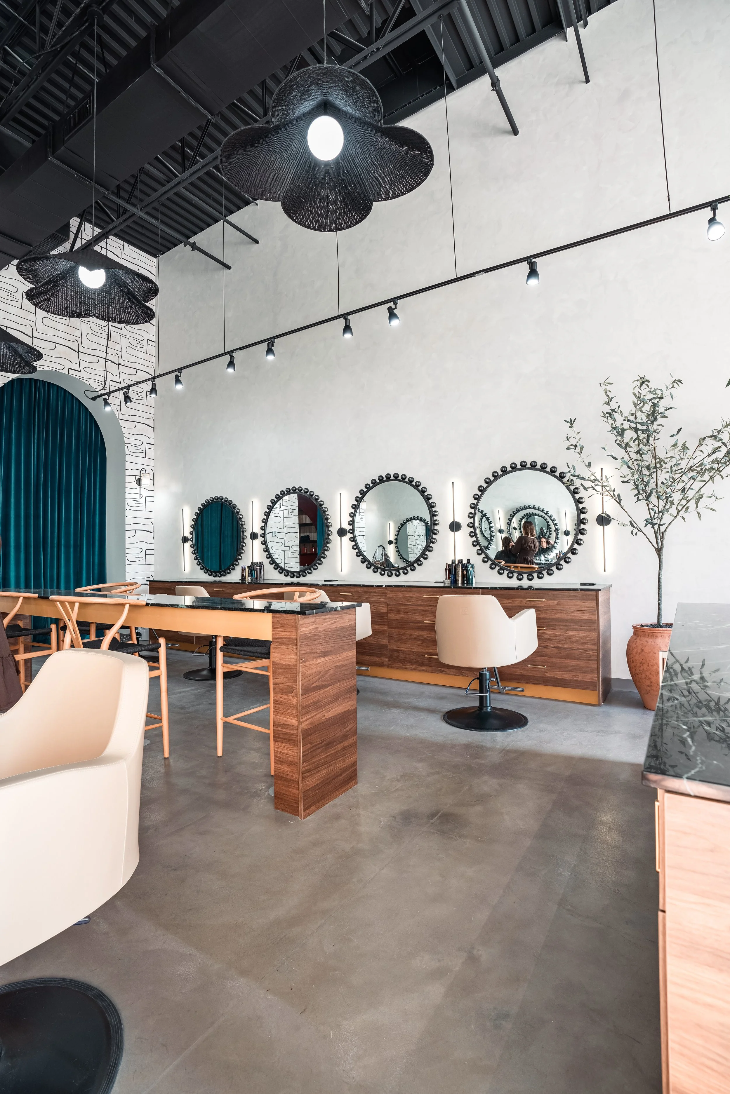 Interior of a modern hair salon with three round mirrors framed with black beads, a wooden styling station, and a beige salon chair, decorated with potted plant and contemporary light fixtures hanging from the ceiling.