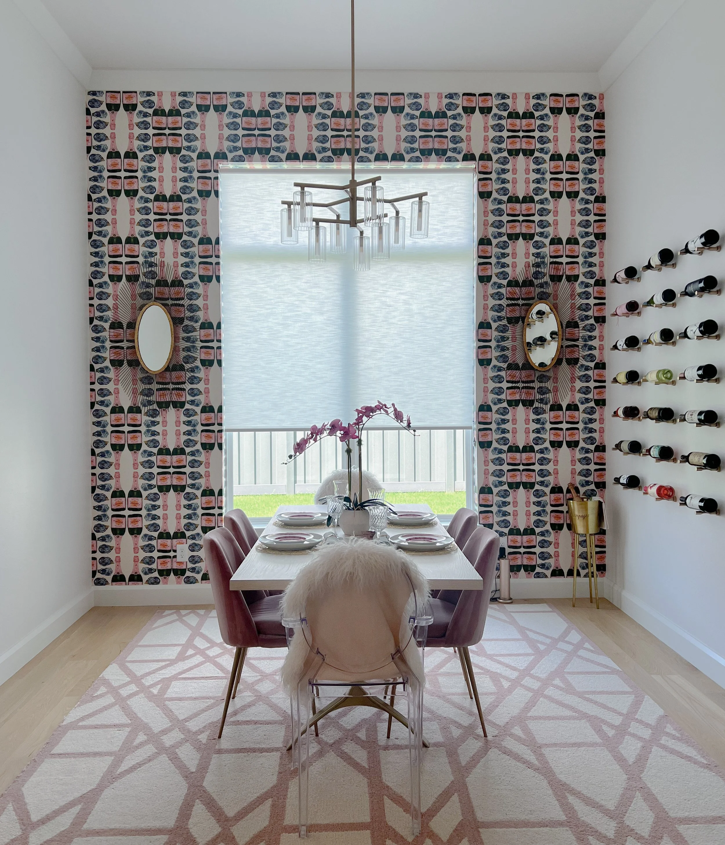 Dining room with a rectangular table set for four, pink velvet chairs, a white furry chair, a pink orchid centerpiece, a pink and white geometric rug, wallpapered accent wall, two oval mirrors, a modern chandelier, window with blinds, and a wall-moun
