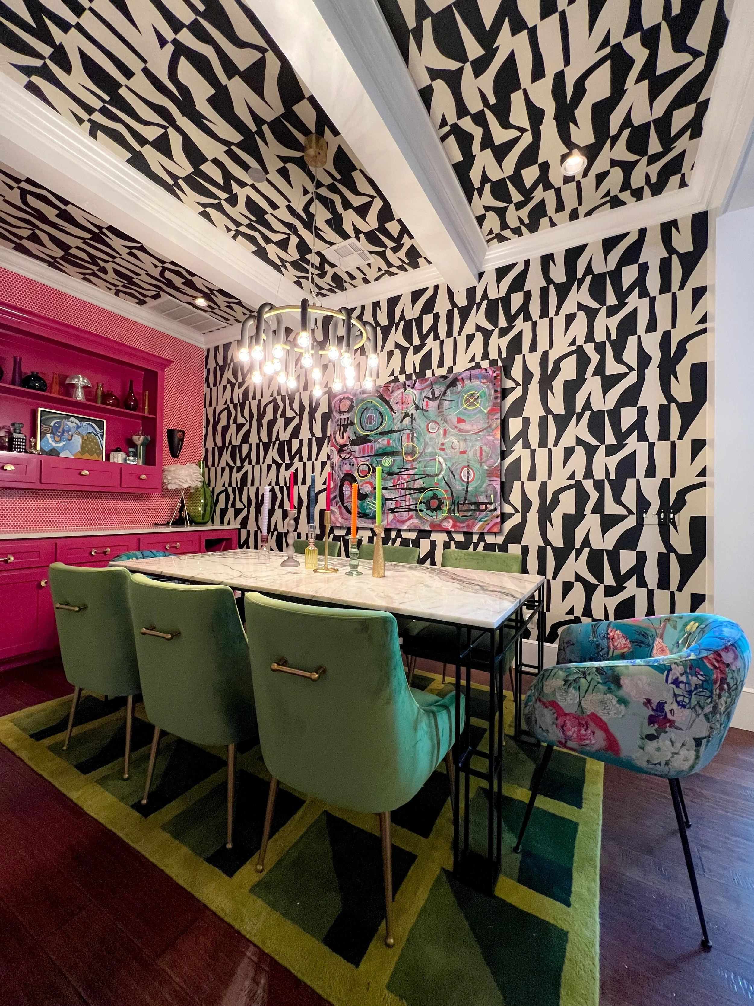 A dining room with black and white abstract patterned wallpaper on the walls and ceiling, a marble dining table, six green upholstered chairs, and an armchair with a floral pattern. A pink cabinet with decorative items and a colorful abstract painting on the wall are also visible. A modern chandelier with exposed bulbs hangs above the table, and the room has a green and black geometric rug on dark wood floors.
