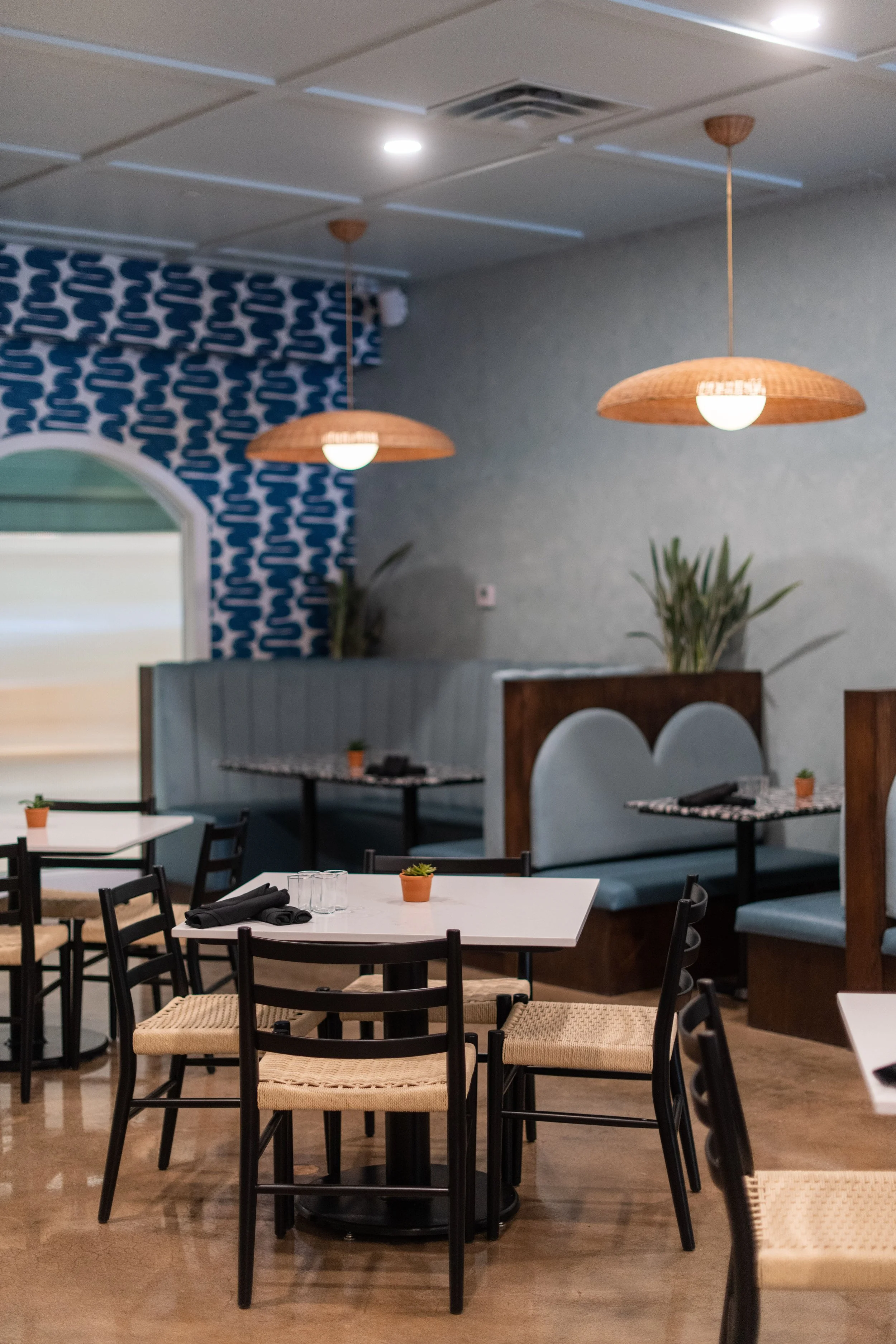 Interior of a restaurant with white tables, black chairs with woven seats, and small potted plants as table centerpieces. Hanging wicker pendant lights and booth seating with blue cushions. Patterned blue and white wallpaper on one wall, plants, and 