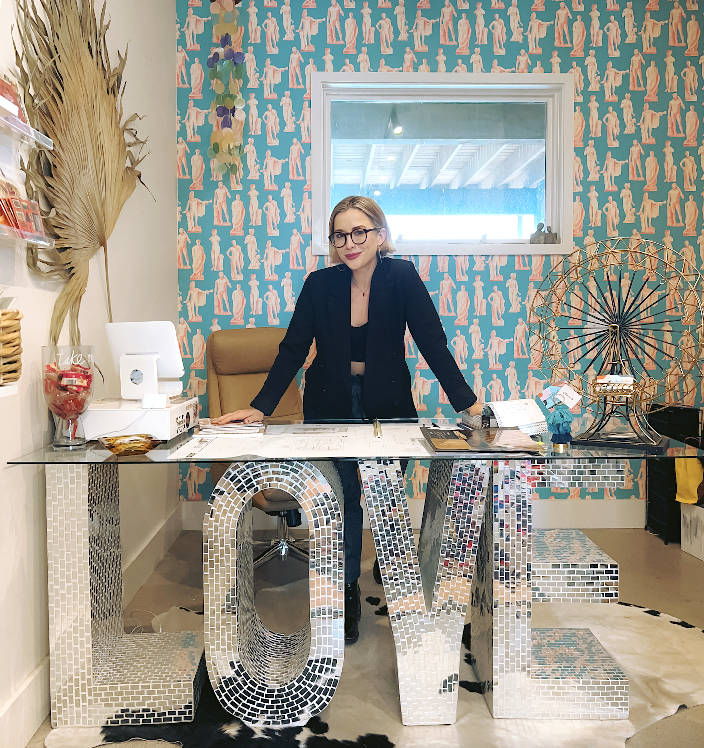 Woman in black blazer and glasses standing behind a desk with large silver 'LOVE' letters decorated with small mirror tiles, in a colorful office space with patterned wallpaper, decorative items, and a window in the background.