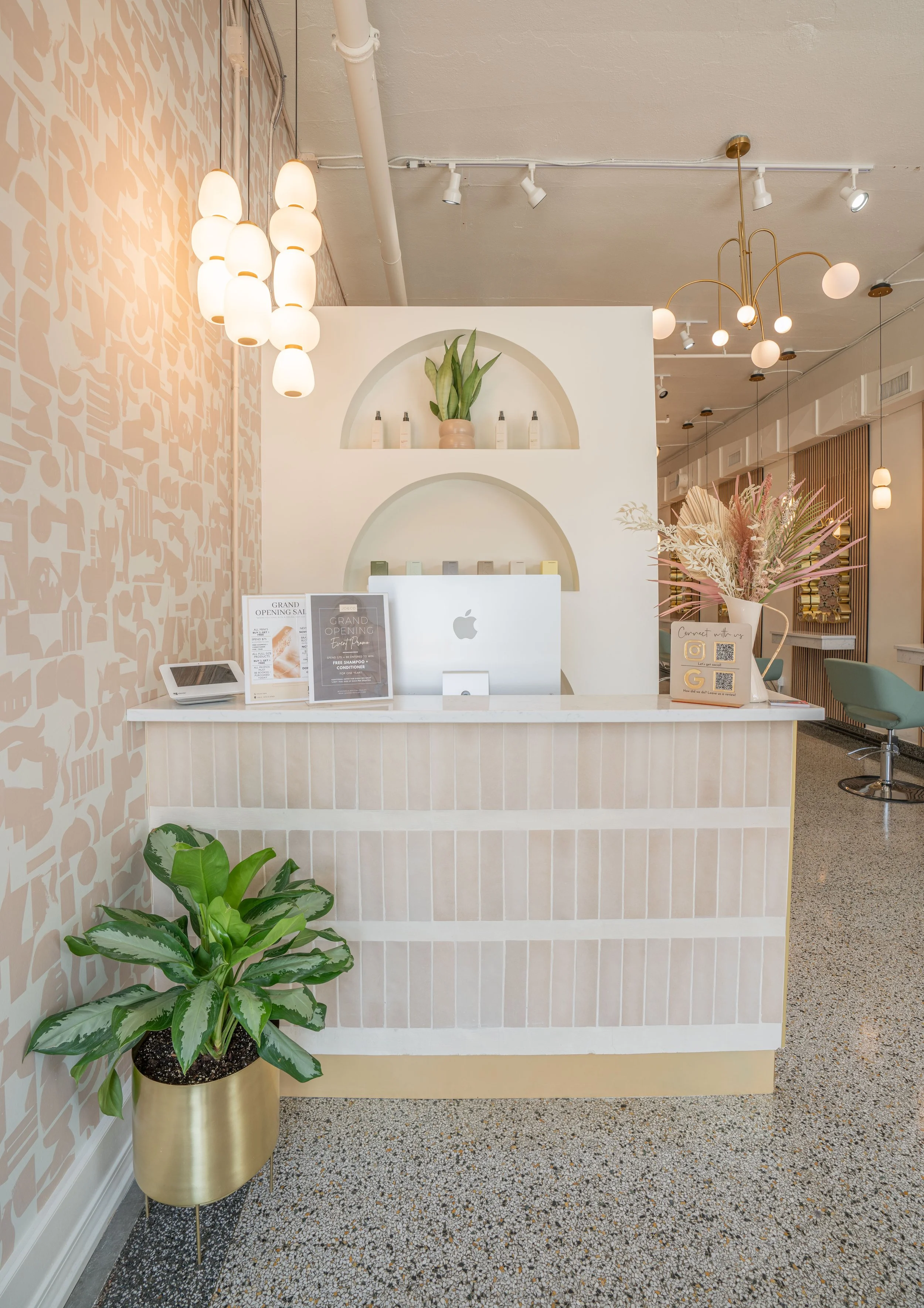 Interior of a modern, minimalist reception area with a white marble counter, green plants, decorative lighting, and pastel-colored chairs