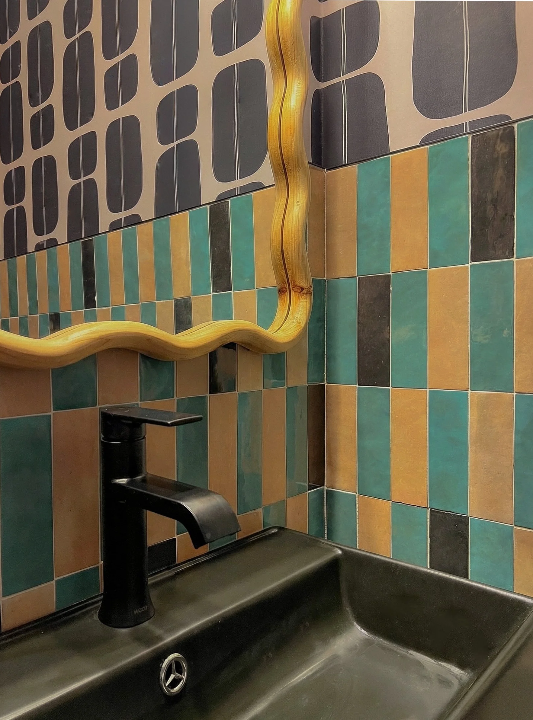 A bathroom sink with a black faucet, a large mirror with a wavy yellow frame, and a wall decorated with multicolored tiles in shades of green, orange, and black, some with unique patterns.