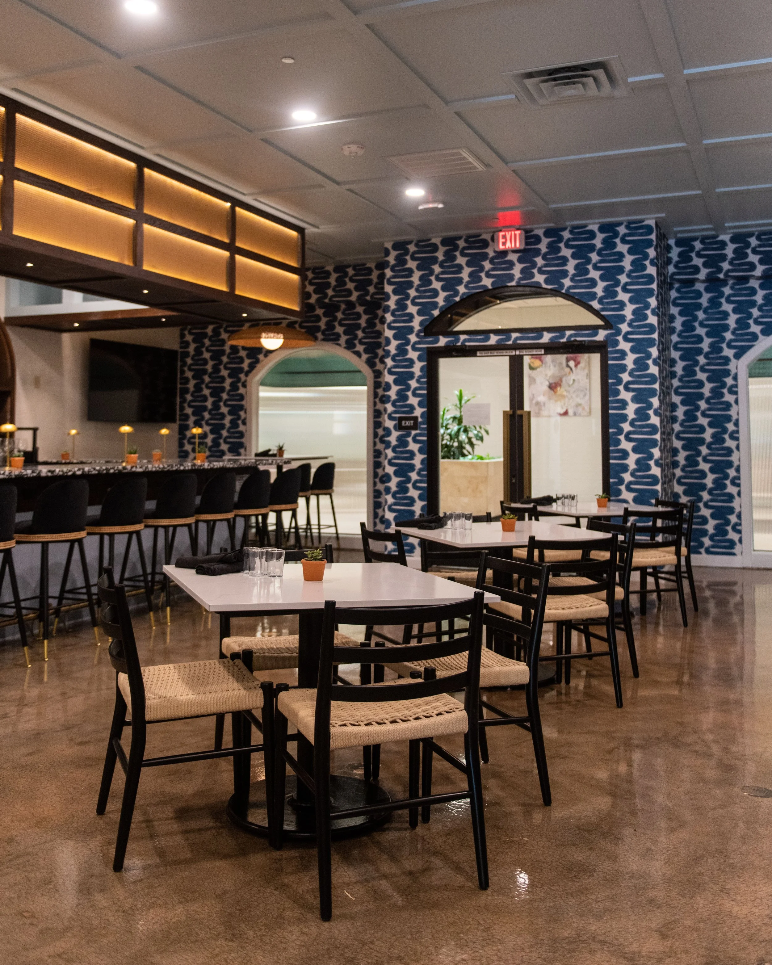 Modern restaurant interior with white tables, black chairs, a bar with high stools, patterned blue and white walls, potted plants, and votive candles.