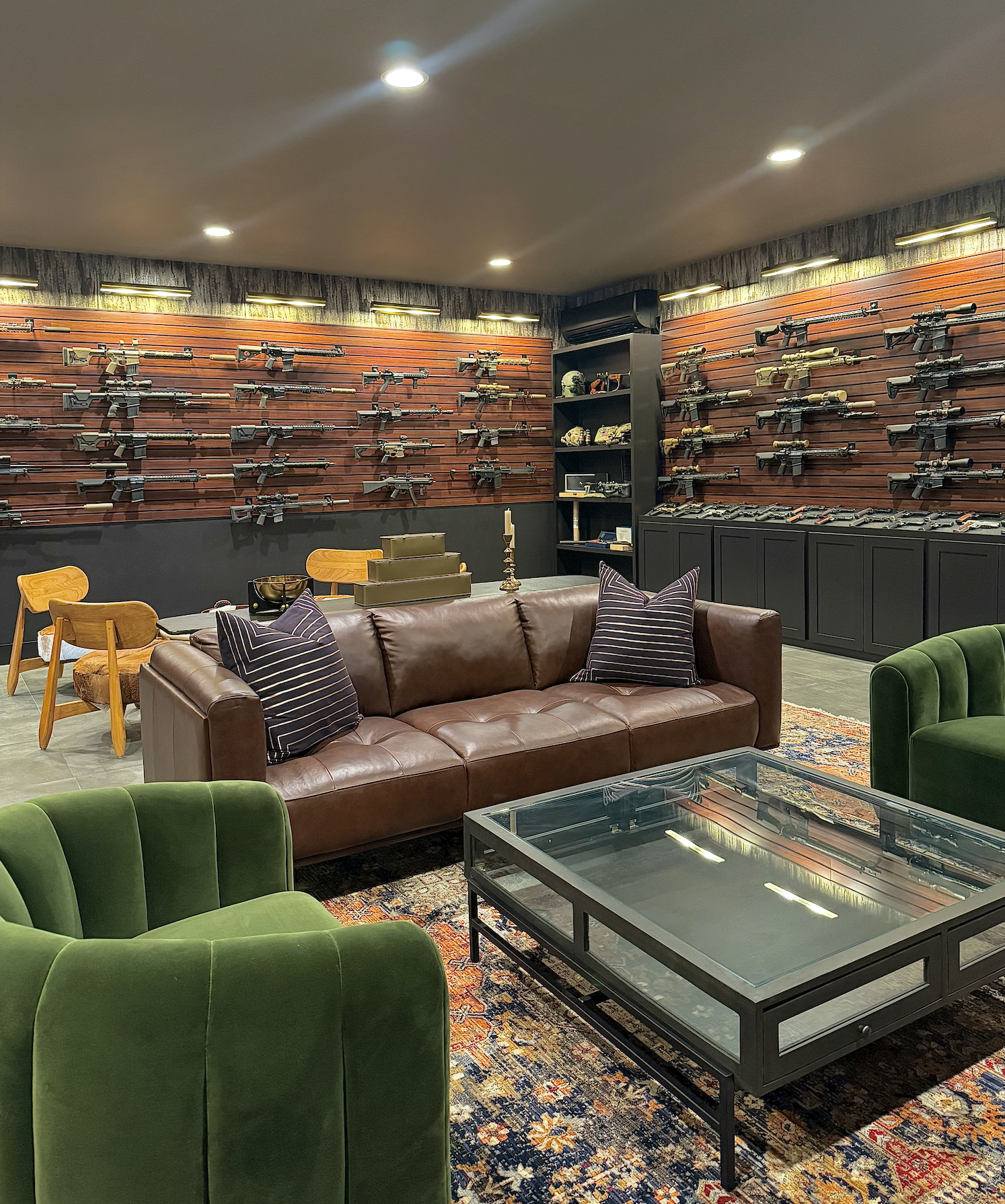 A room decorated like a game or shooting lounge with a wall of guns displayed on a wooden backdrop, a leather sofa, green velvet chairs, a glass coffee table, and a dining area with chairs.