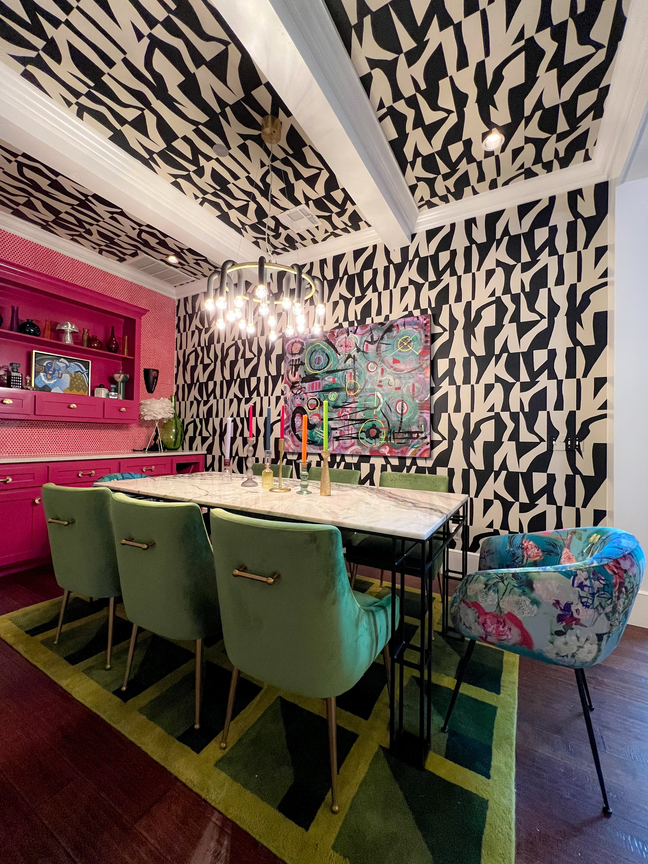 A dining room with black and white abstract patterned wallpaper on the walls and ceiling, a white marble table with green chairs, a floral upholstered armchair, and a pink cabinet with decorative items on the left. There is a modern chandelier hangin