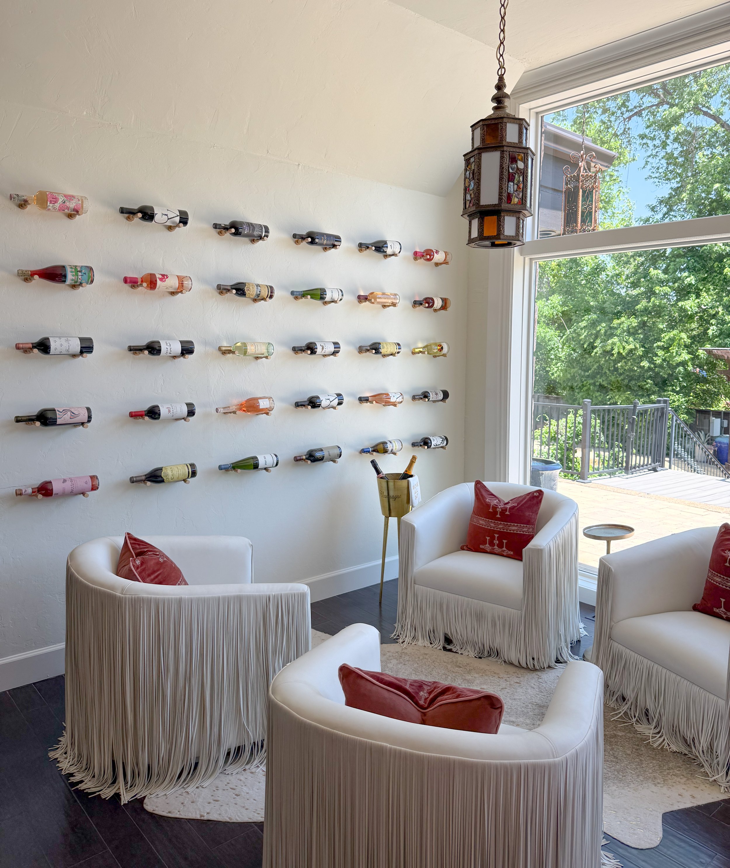 A living room with four white chairs with fringed skirts and red pillows, a small side table, large window with a view of the outdoors, and a wall-mounted wine rack with multiple bottles, with a decorative lantern hanging from the ceiling.