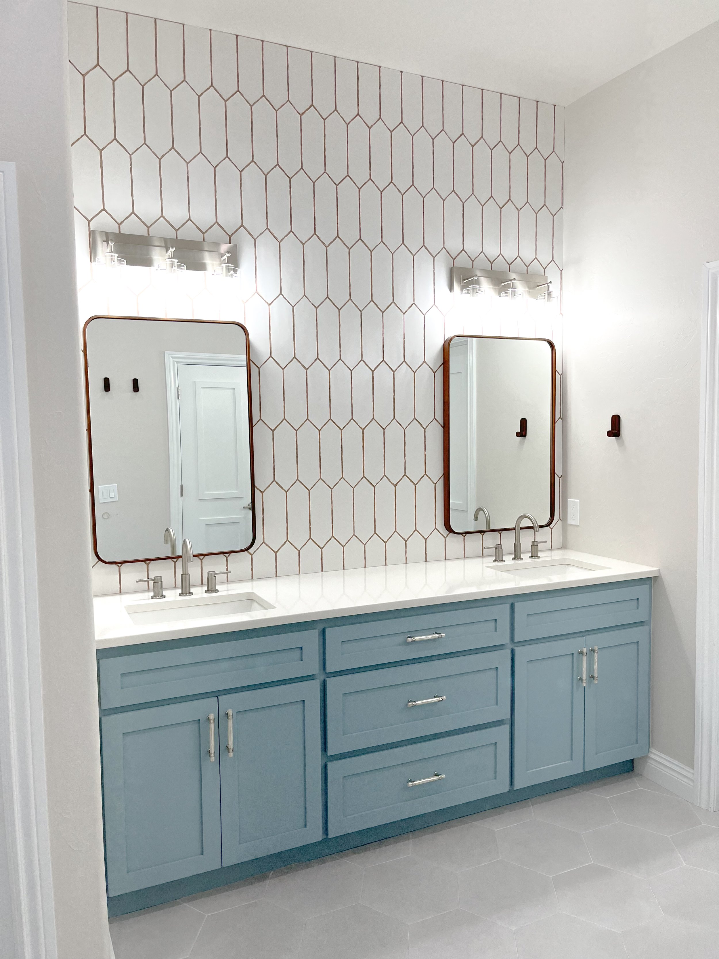Double bathroom vanity with blue cabinets, white countertop, two rectangular mirrors with rounded edges, and modern light fixtures above each mirror. The wall behind the mirrors features a honeycomb pattern tile design.