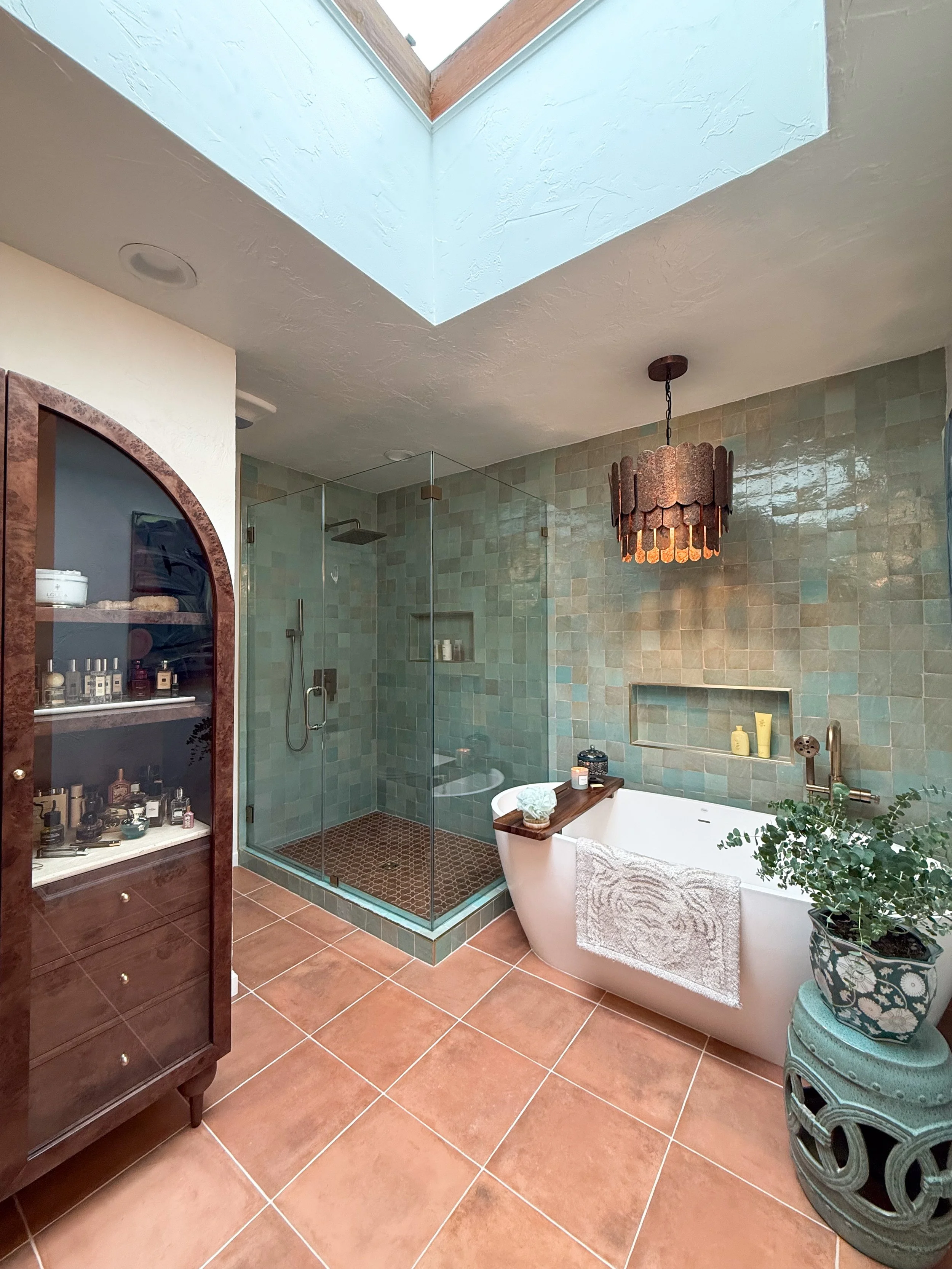 Bathroom featuring a glass-enclosed shower with green tiles, a freestanding bathtub, a wooden shelf on the bathtub with toiletries, a hanging brown chandelier, a wooden cabinet with toiletries, and a potted plant on an ornate stool.