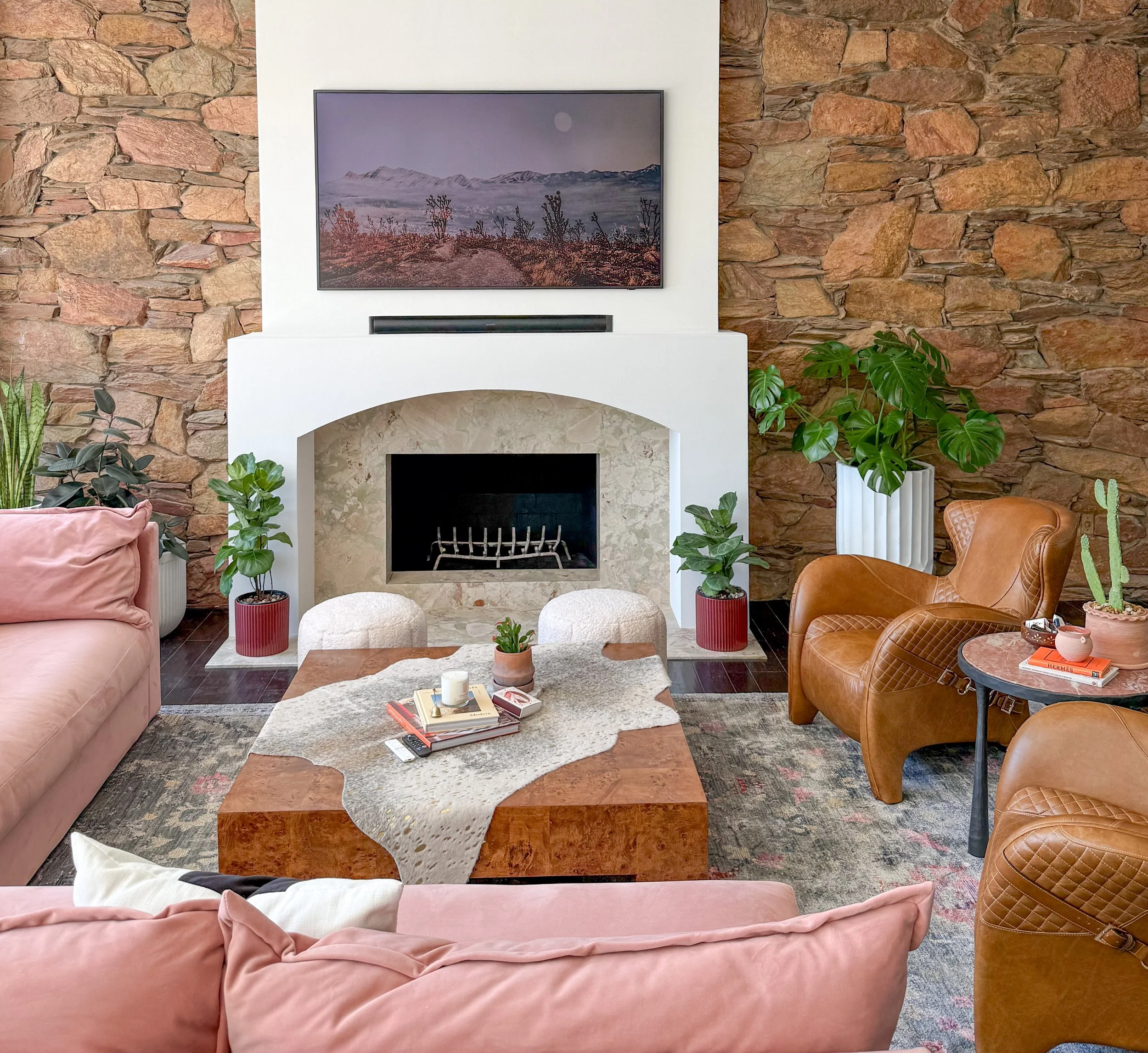 Living room with a stone accent wall, a fireplace, a flat-screen TV above the fireplace, a pink sofa, brown leather chairs, a wooden coffee table, and various green potted plants.