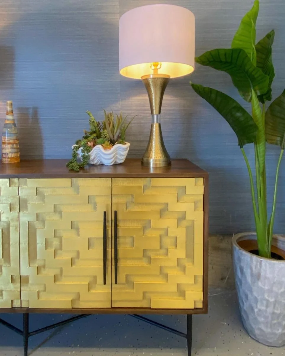 A modern wooden sideboard with a yellow textured front, black handles, and black legs, topped with plants, a lamp with a gold and silver base and white shade, and a decorative bottle, situated in a room with gray textured walls and a large potted gre