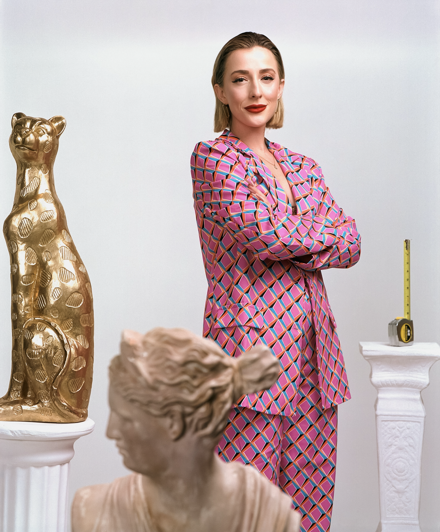 A woman with blonde hair, wearing a colorful geometric patterned suit, stands with arms crossed in front of a white wall, surrounded by classical sculptures and a measuring tape on a pedestal.