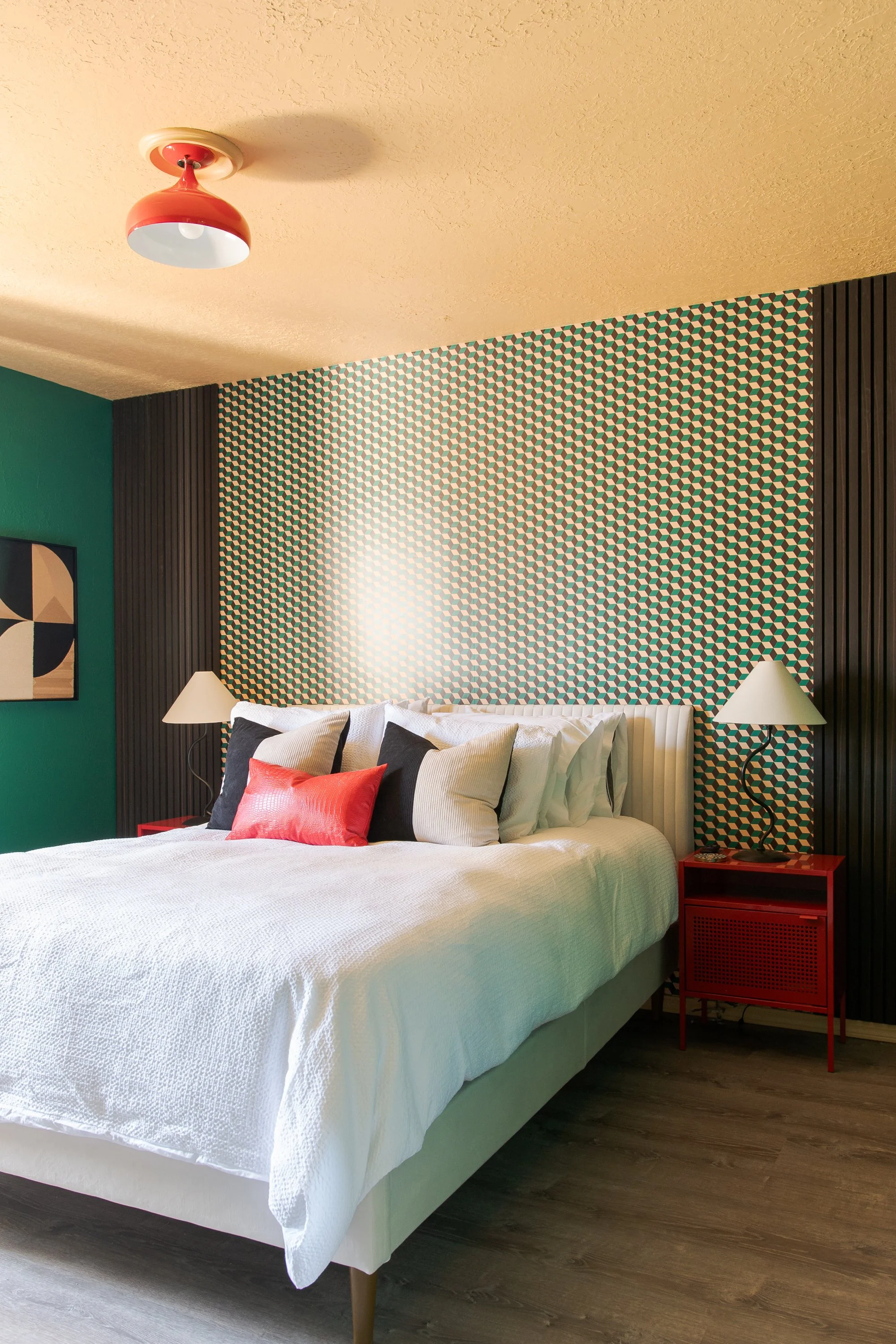 A modern bedroom with a bed, multiple pillows, a red bedside table, and two lamps with white shades on black curving bases. The wall behind the bed has geometric patterned wallpaper, flanked by black vertical paneling; another wall is painted green w