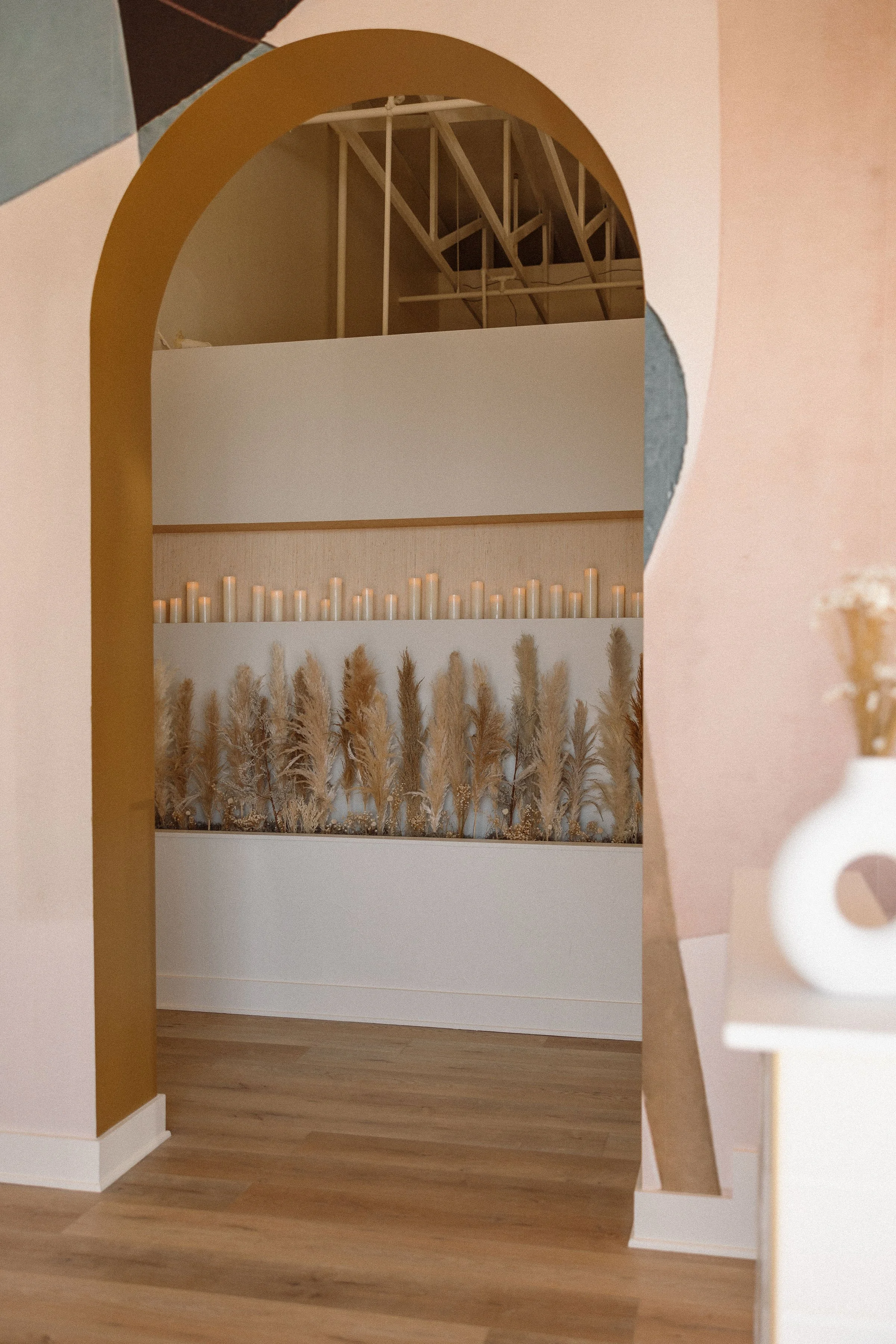 An interior space viewed through an archway, featuring a wall with a row of lit candles, decorative dried plants, and a wooden floor.
