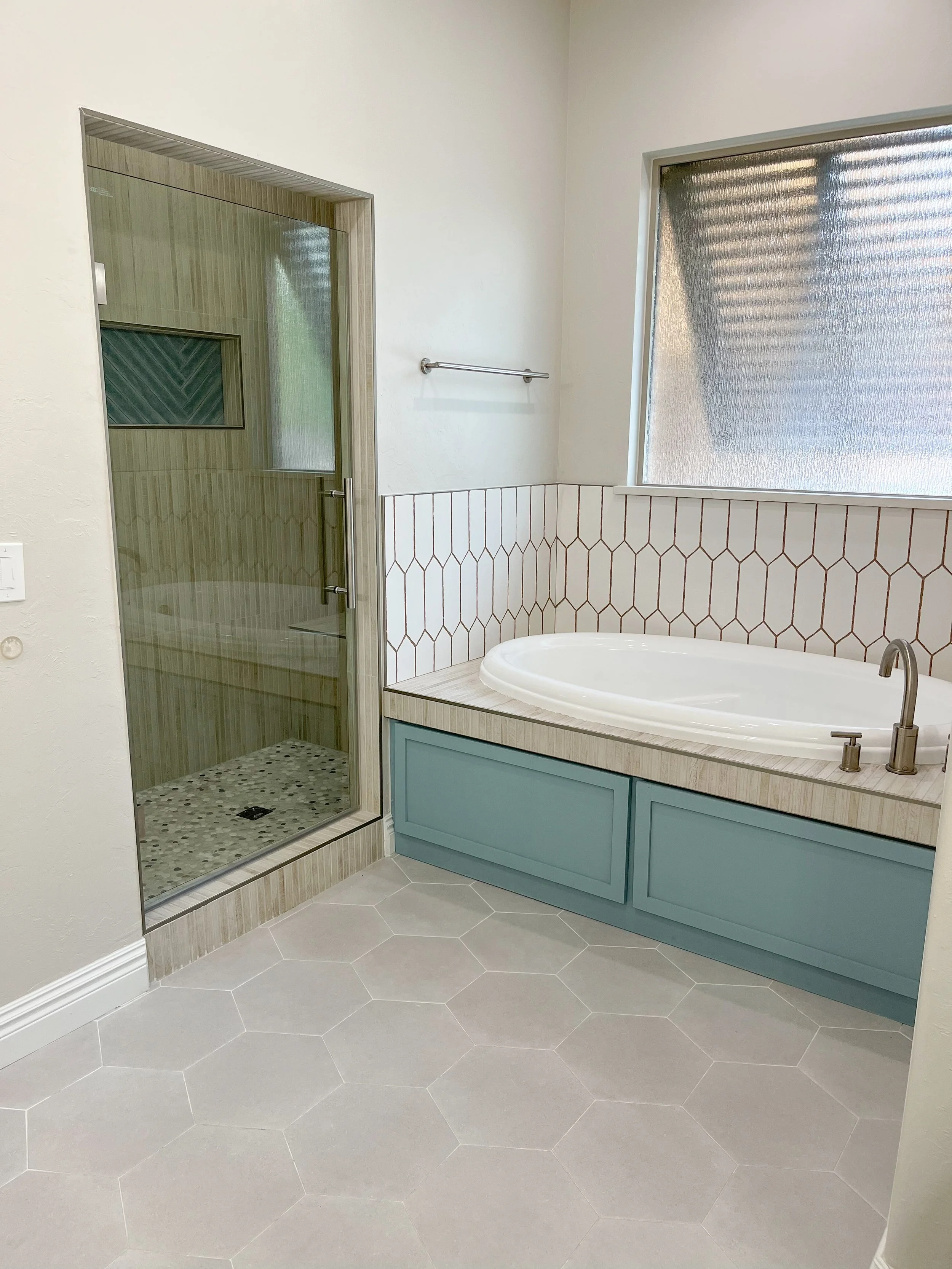 Bathroom with a glass shower enclosure, a white bathtub with a light blue wooden cabinet, a large frosted window, and hexagonal floor tiles.