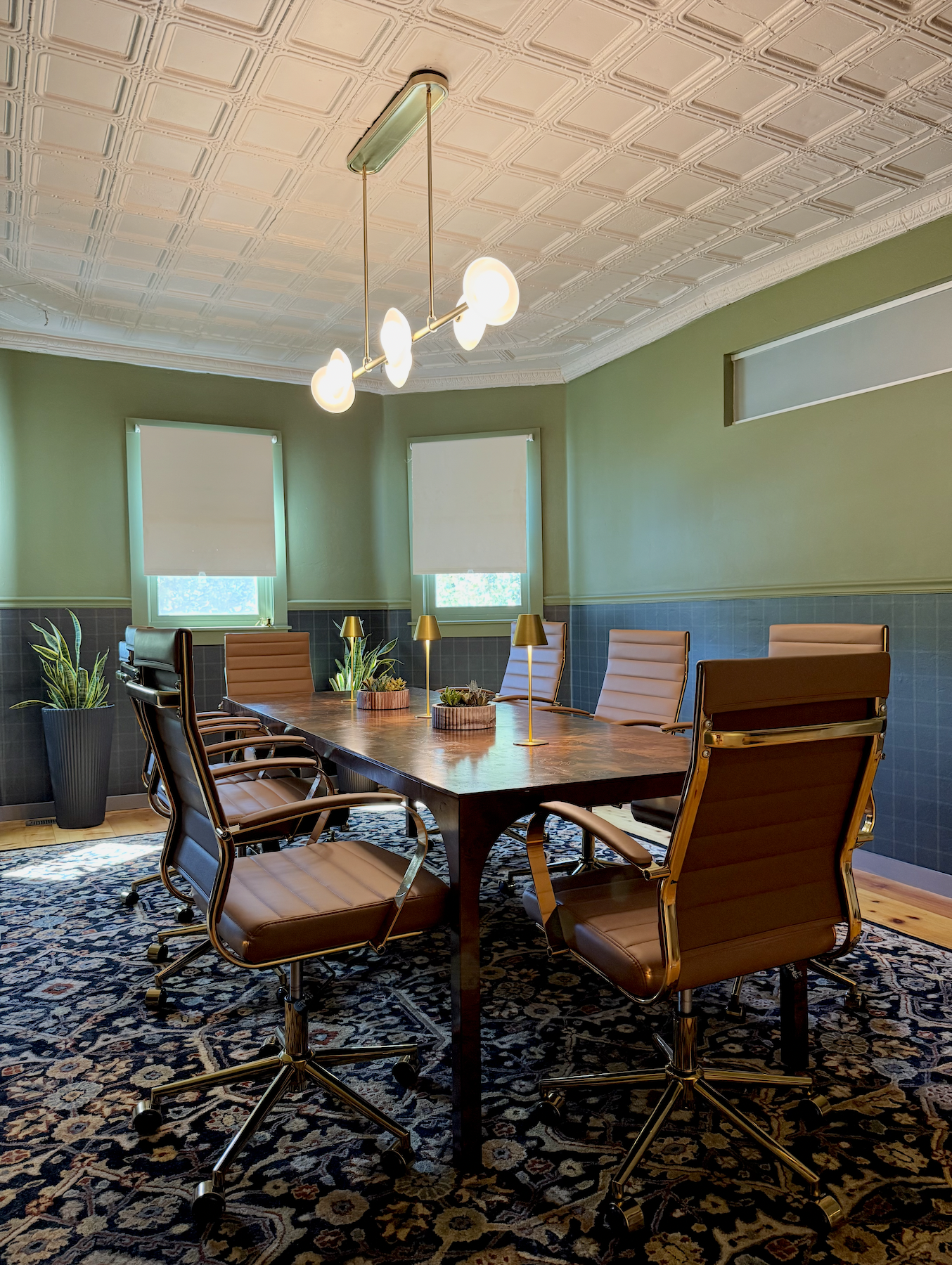 Empty conference room with a wooden table, eight brown leather office chairs, a patterned rug, three potted plants, two window blinds, and a modern ceiling light fixture.