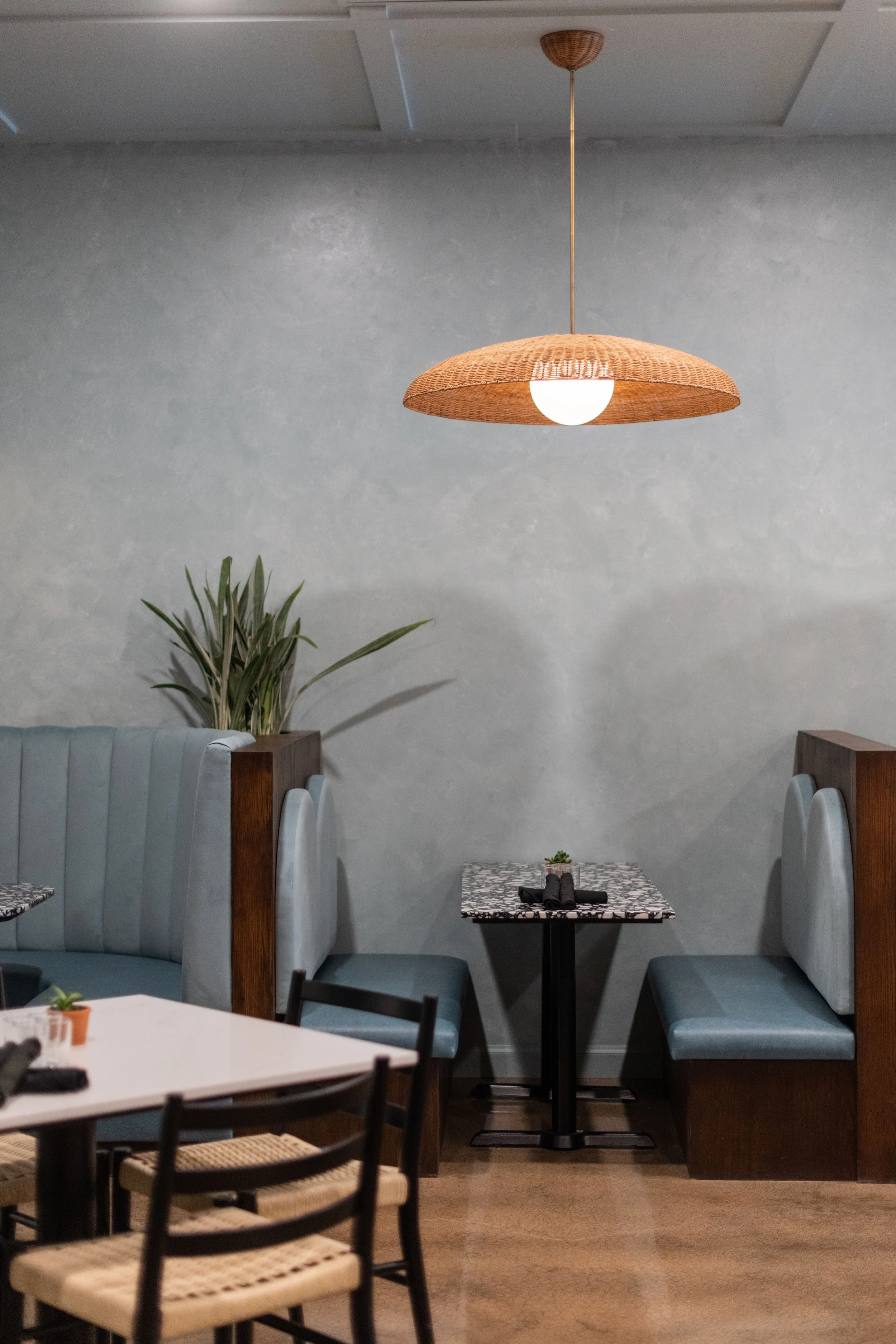 A cozy restaurant corner featuring a hanging wicker lampshade with a white globe bulb, blue upholstered booth seating with high backs, a small marble-topped table with black utensils and a small potted plant, and a large indoor plant with long leaves