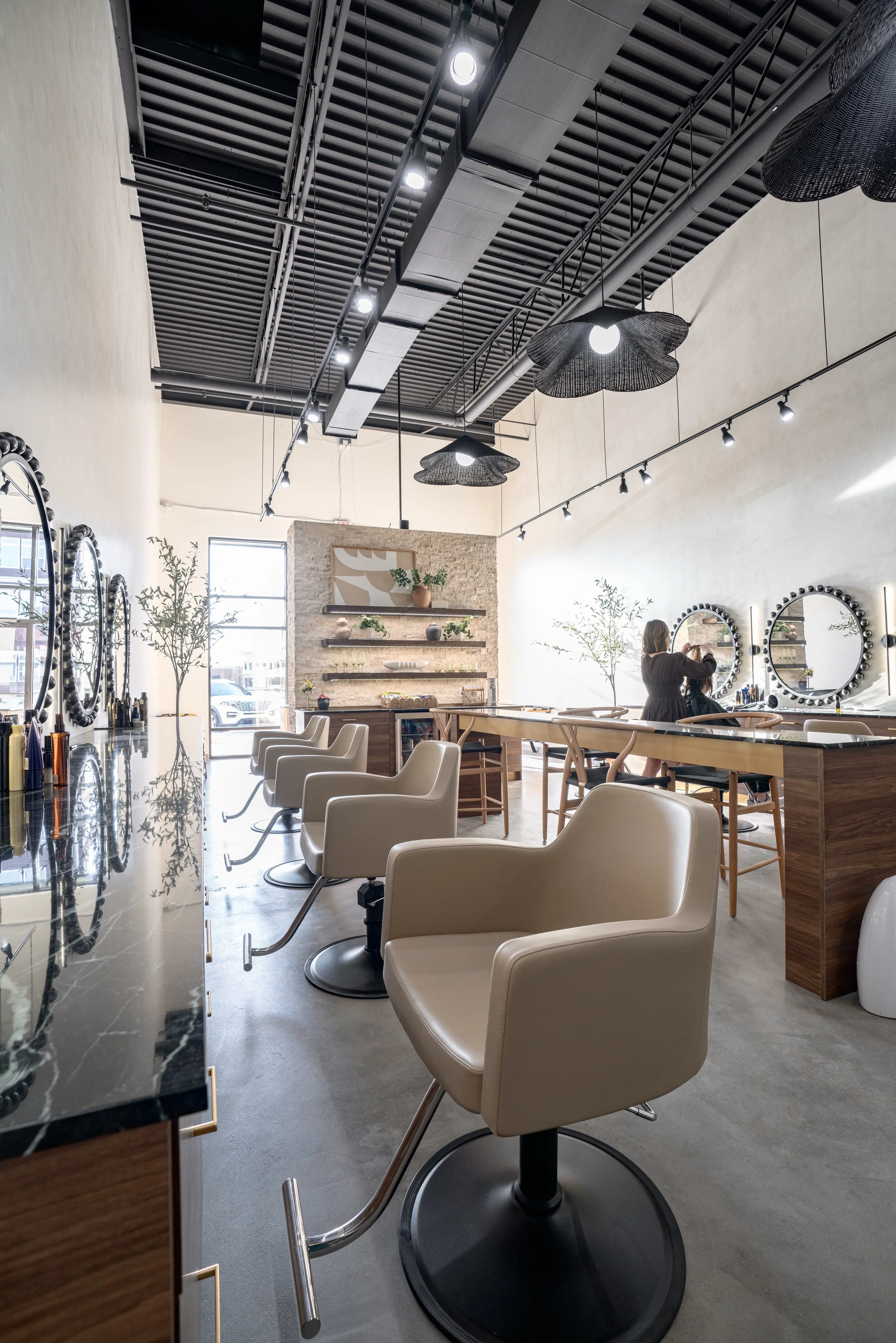 Interior of a modern salon with beige chairs, mirrors with black decorative frames, and a woman at a sink. The space has high ceilings with black industrial-style decor and hanging black flower-shaped pendant lights, natural light from large windows,