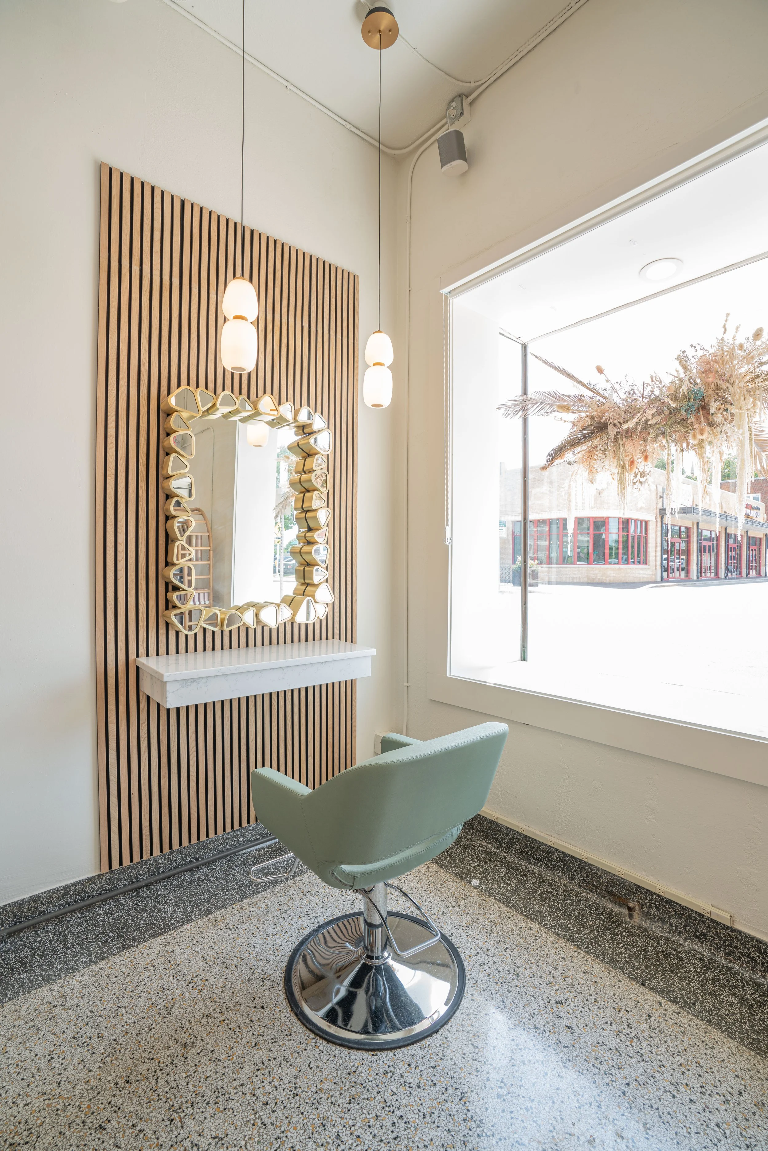 Interior of a modern salon with a mint green salon chair, wooden slat wall with decorative mirror, white marble shelf, and hanging pendant lights beside a large window showing an outdoor view with dried flower arrangement.
