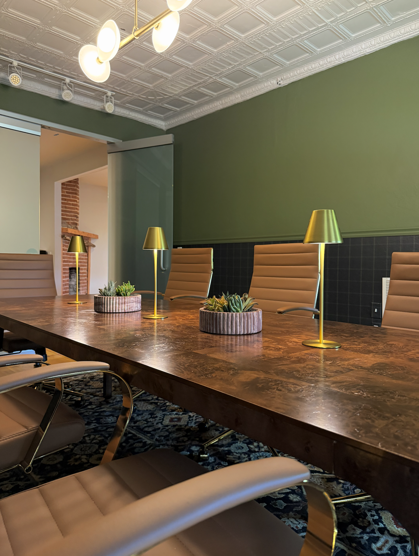 A conference room with a large wooden table, tan leather chairs, gold desk lamps, and small potted succulents. The room has a green wall, a decorative ceiling, and a patterned rug.