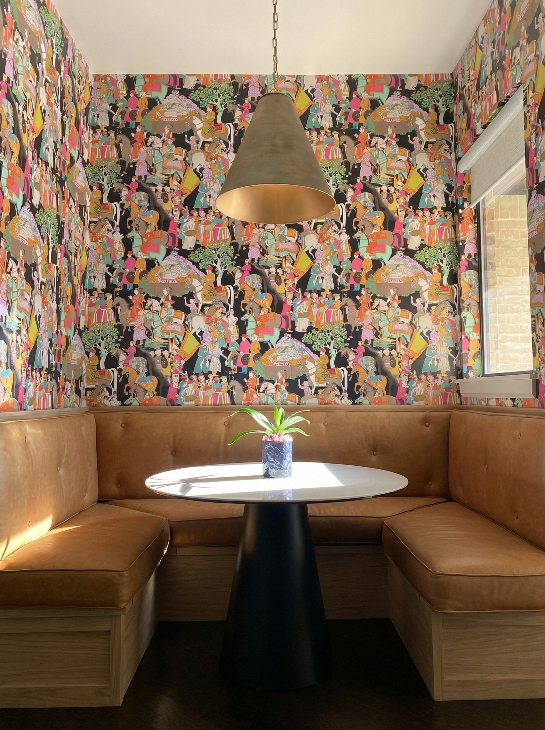 A cozy dining nook with brown leather banquette seating around a white round table, decorated with a small potted plant. The backdrop features colorful, patterned wallpaper with various figures and scenes, a brass pendant lamp hanging from the ceilin