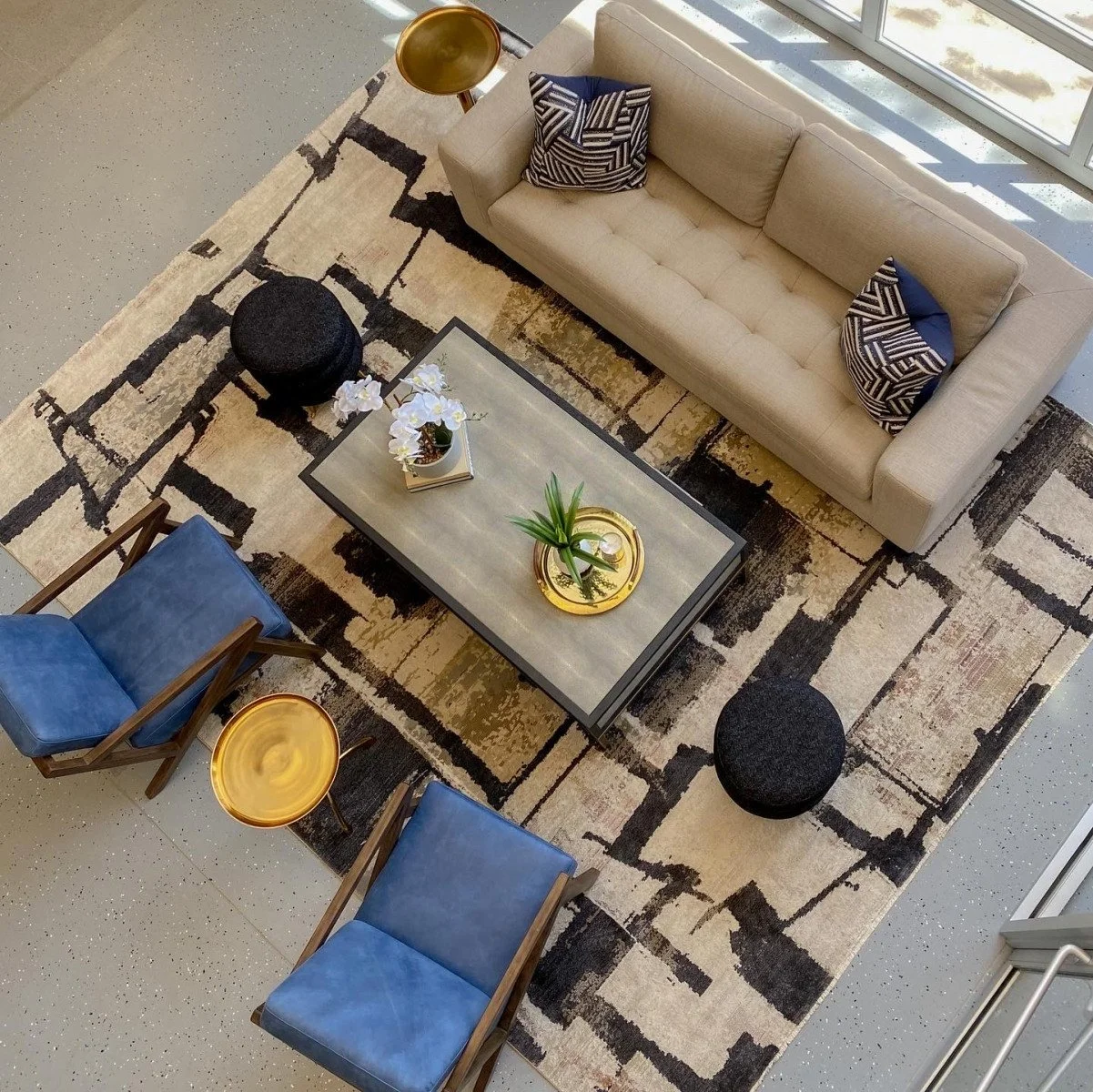 A top-down view of a modern living room with a beige sofa, two blue chairs, and a glass coffee table with decorative items. A patterned rug covers the floor, and large windows provide natural light.