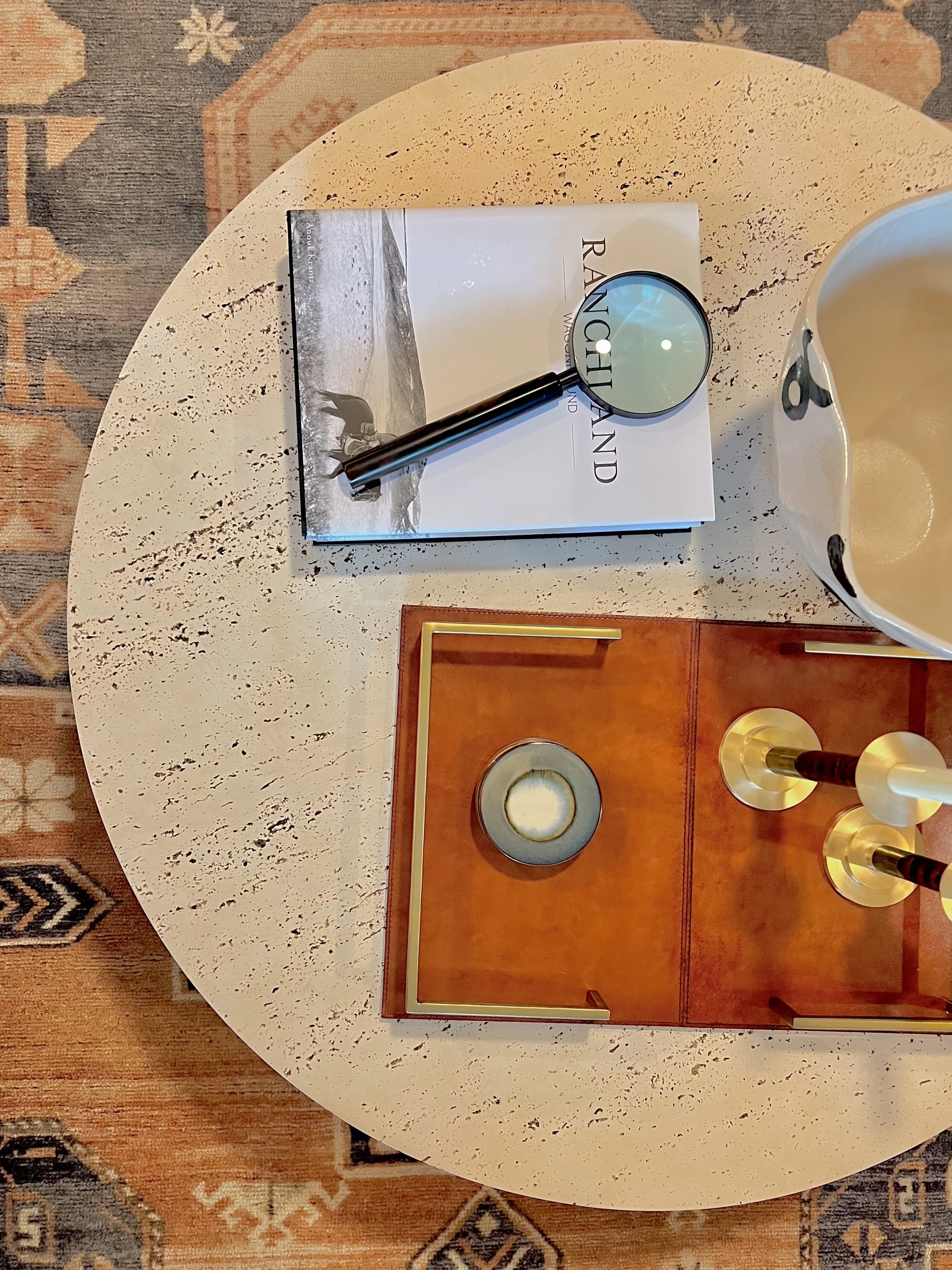 A round white table with a book titled 'Ranchland' and a magnifying glass on it, along with decorative items including a wooden tray with candles and a large white and black ceramic bowl, all placed on a patterned rug.