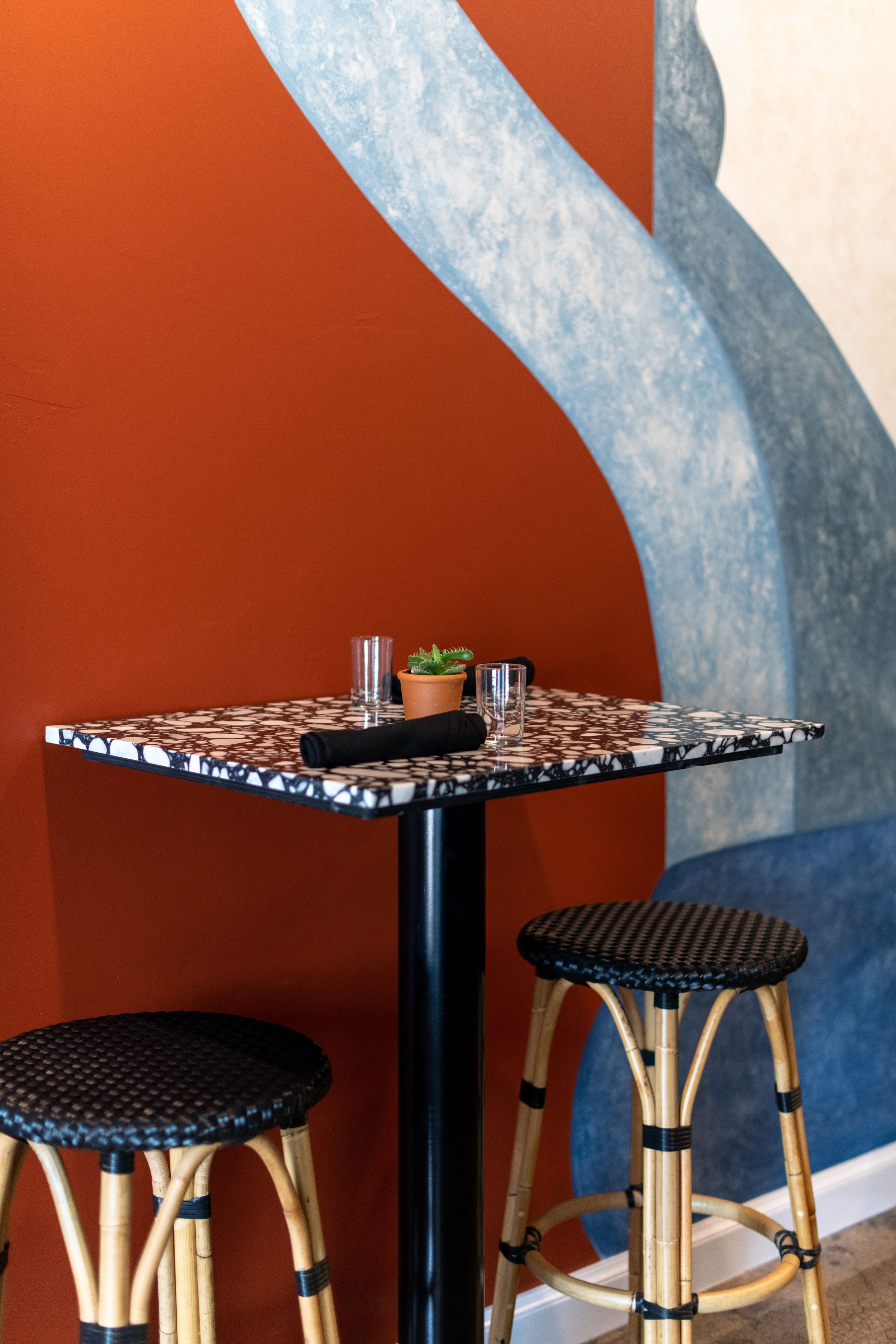 A small table with a black and white patterned top, set with a potted succulent, two glasses, and a napkin, positioned against a colorful abstract wall with blue, orange, and cream sections, accompanied by two rattan barstools with woven black seats.
