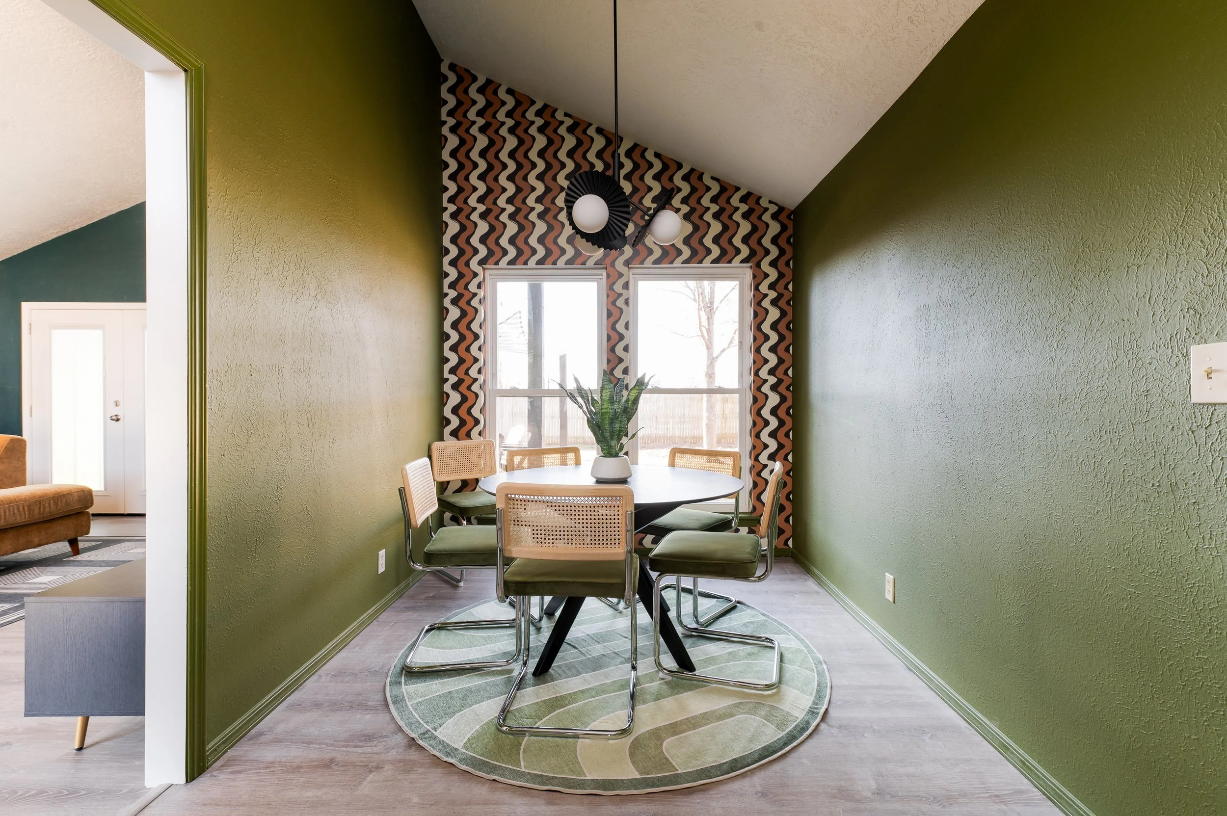 Dining area with green textured walls, a round table with six chairs, potted plant, patterned wallpaper behind the windows, pendant light, and a green circular rug on hardwood floor.