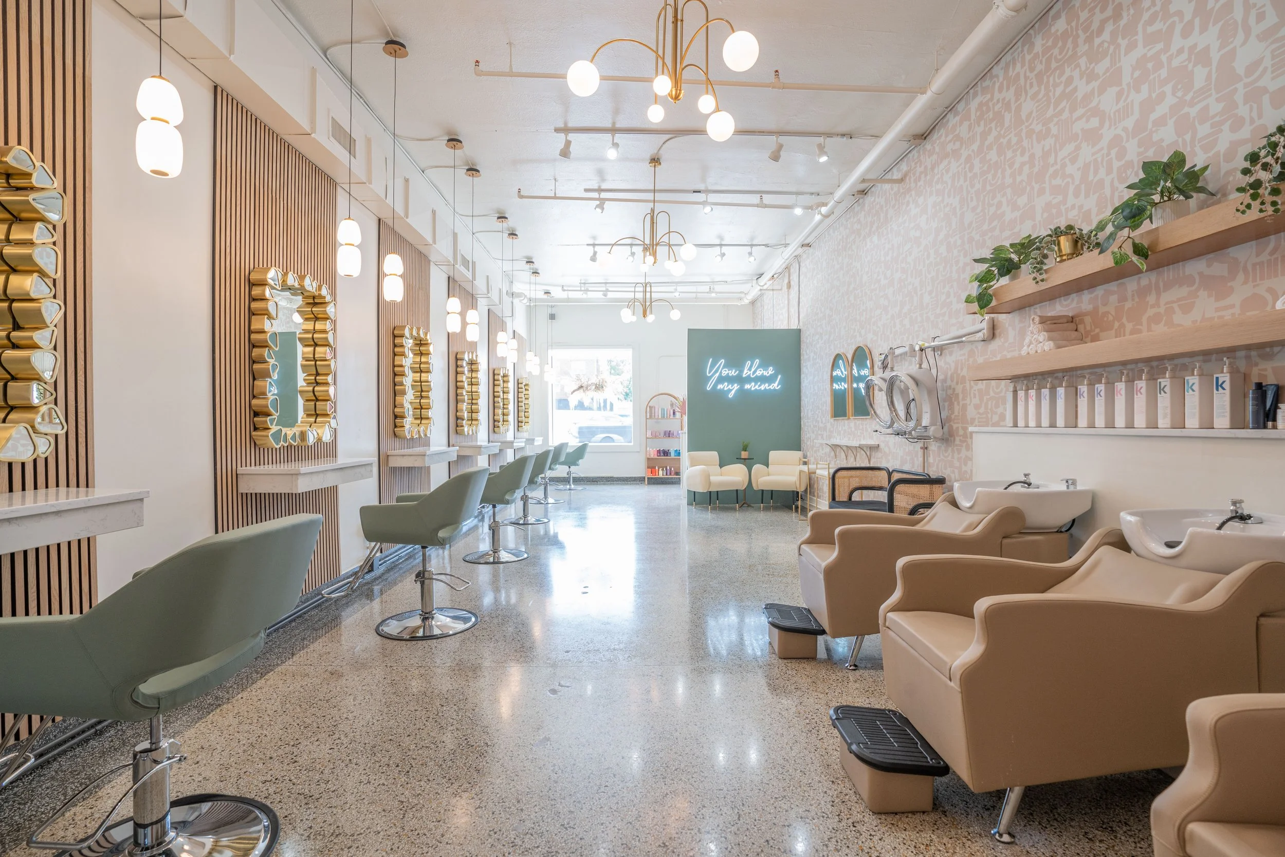 Interior of a modern salon with styling stations and mirrors on the left, and washing stations with sinks and chairs on the right. The decor features soft colors, wooden accents, hanging light fixtures, and a neon sign on a teal wall that says "You b