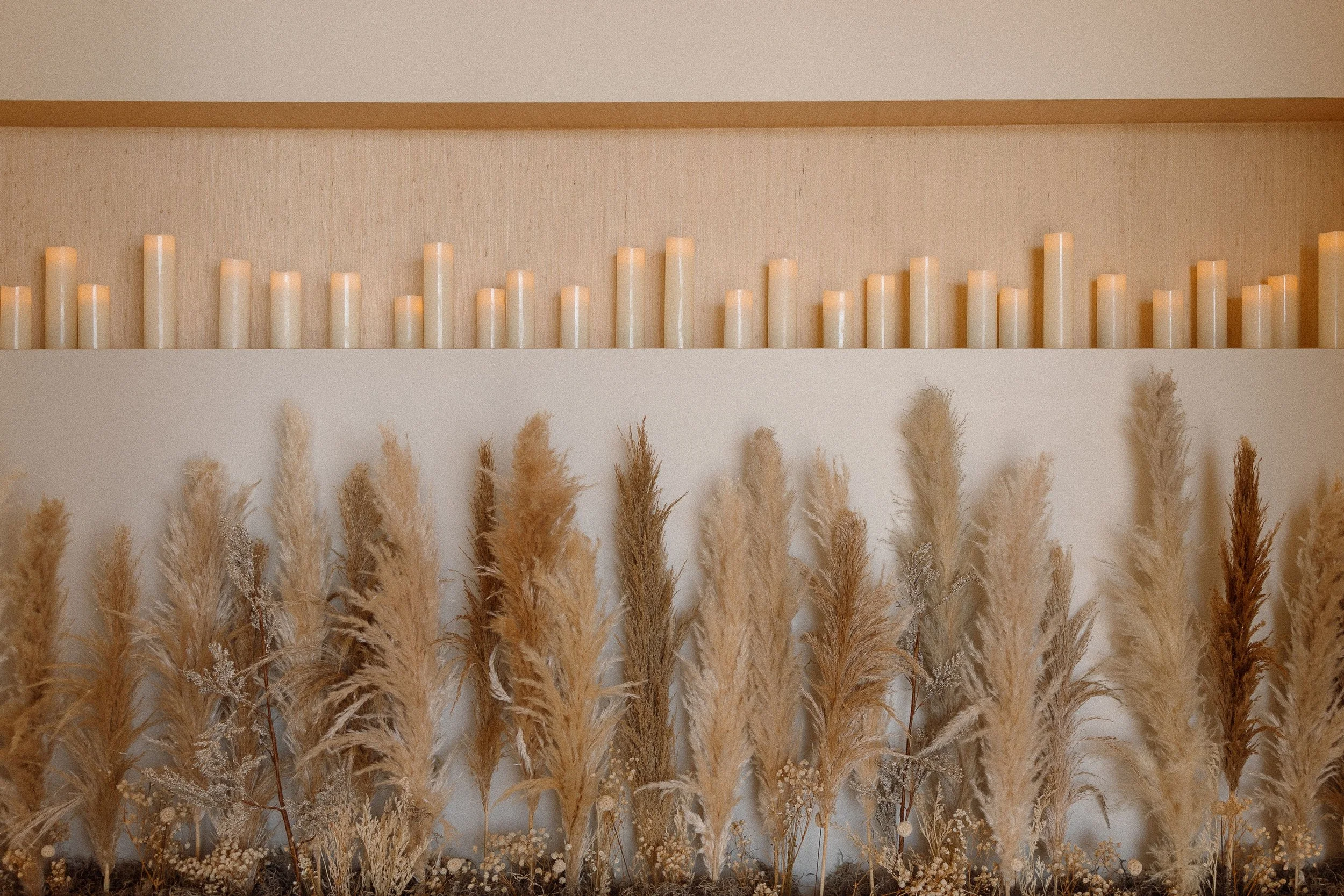 A shelf with white candles at the top and a row of dried pampas grass below, arranged in front of a light beige wall.