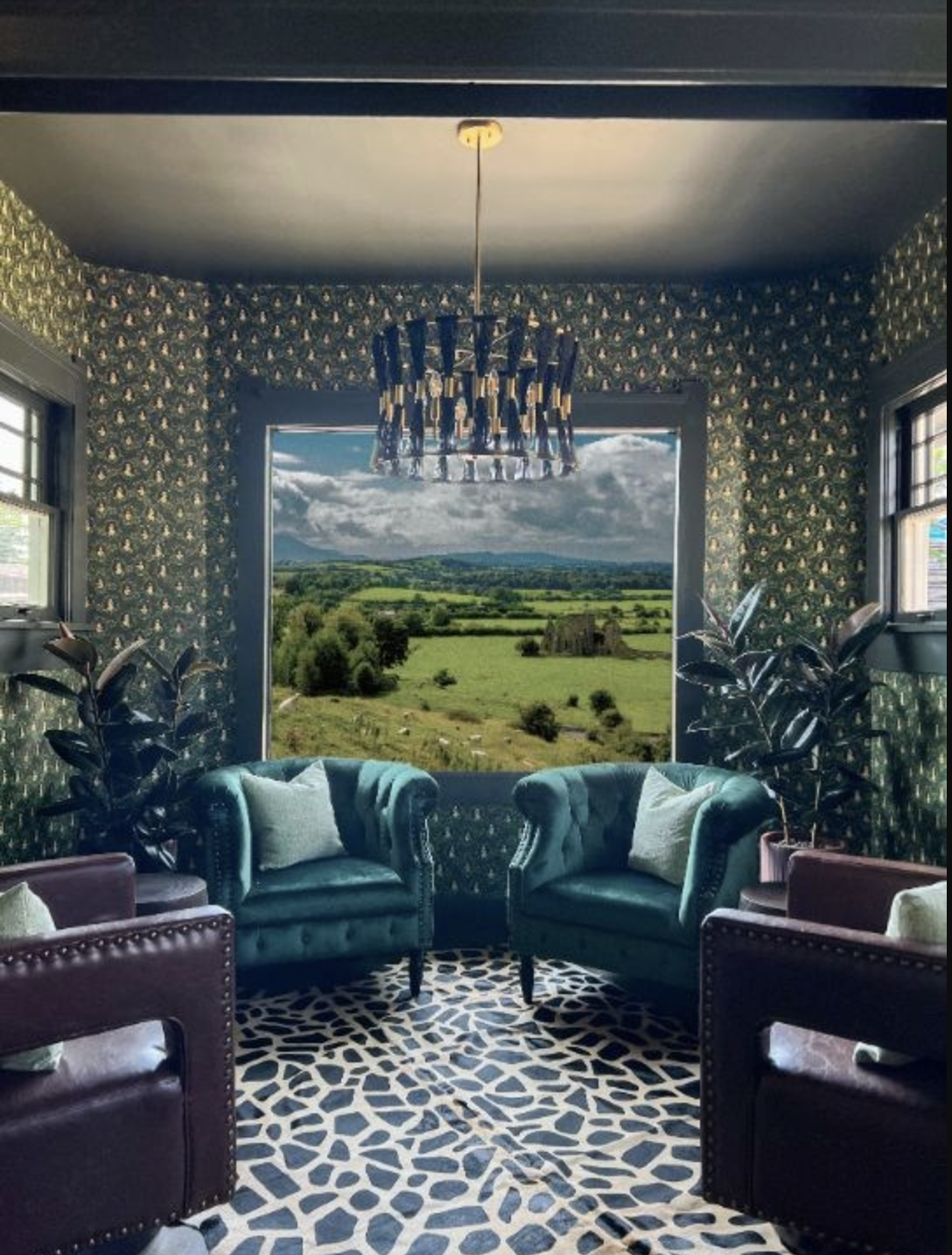Interior of a room with patterned wallpaper, two turquoise velvet armchairs with pillows, and a large window showing a countryside landscape with green fields, trees, and distant mountains under a cloudy sky.