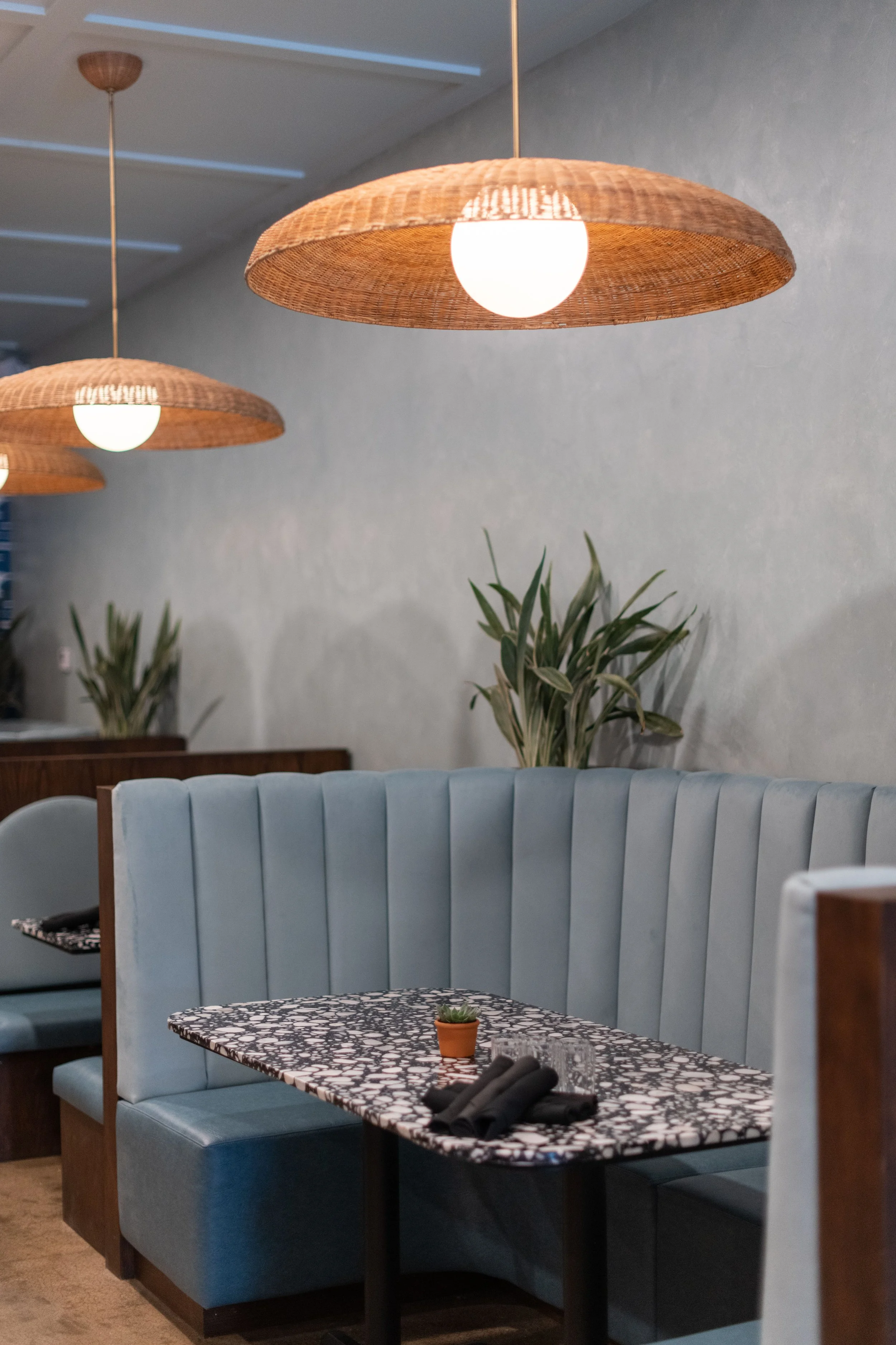 Interior of a restaurant with blue upholstered booths, a marble patterned table set with black napkins, a small potted plant, and two hanging rattan lamps.