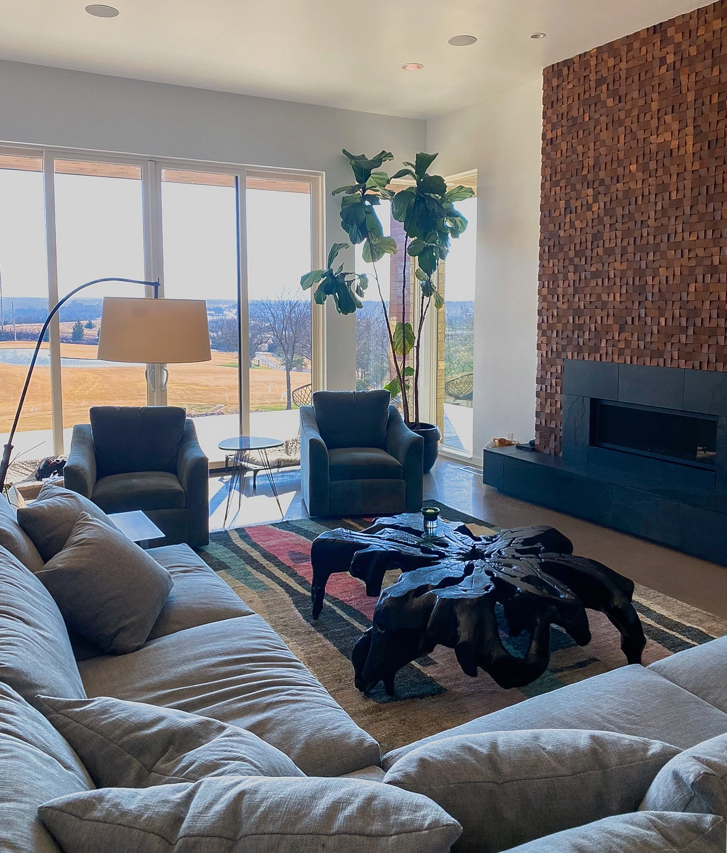 Living room with large windows showing an outdoor landscape, with seating including a sectional sofa and two armchairs, a black coffee table, a tall potted plant, a floor lamp, and a fireplace with a textured wooden wall.