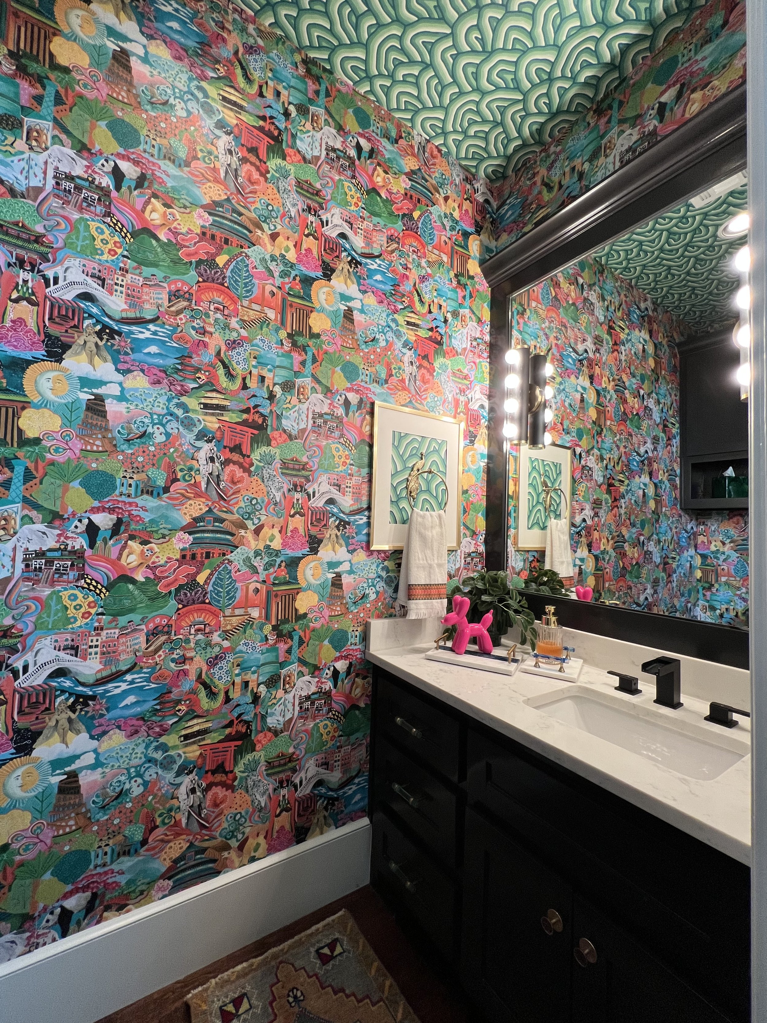 A colorful bathroom with vibrant patterned wallpaper depicting various scenes and structures. A large mirror with lights, a black vanity with a white marble top, and decor items including a pink dog-shaped sculpture and a plant are visible.