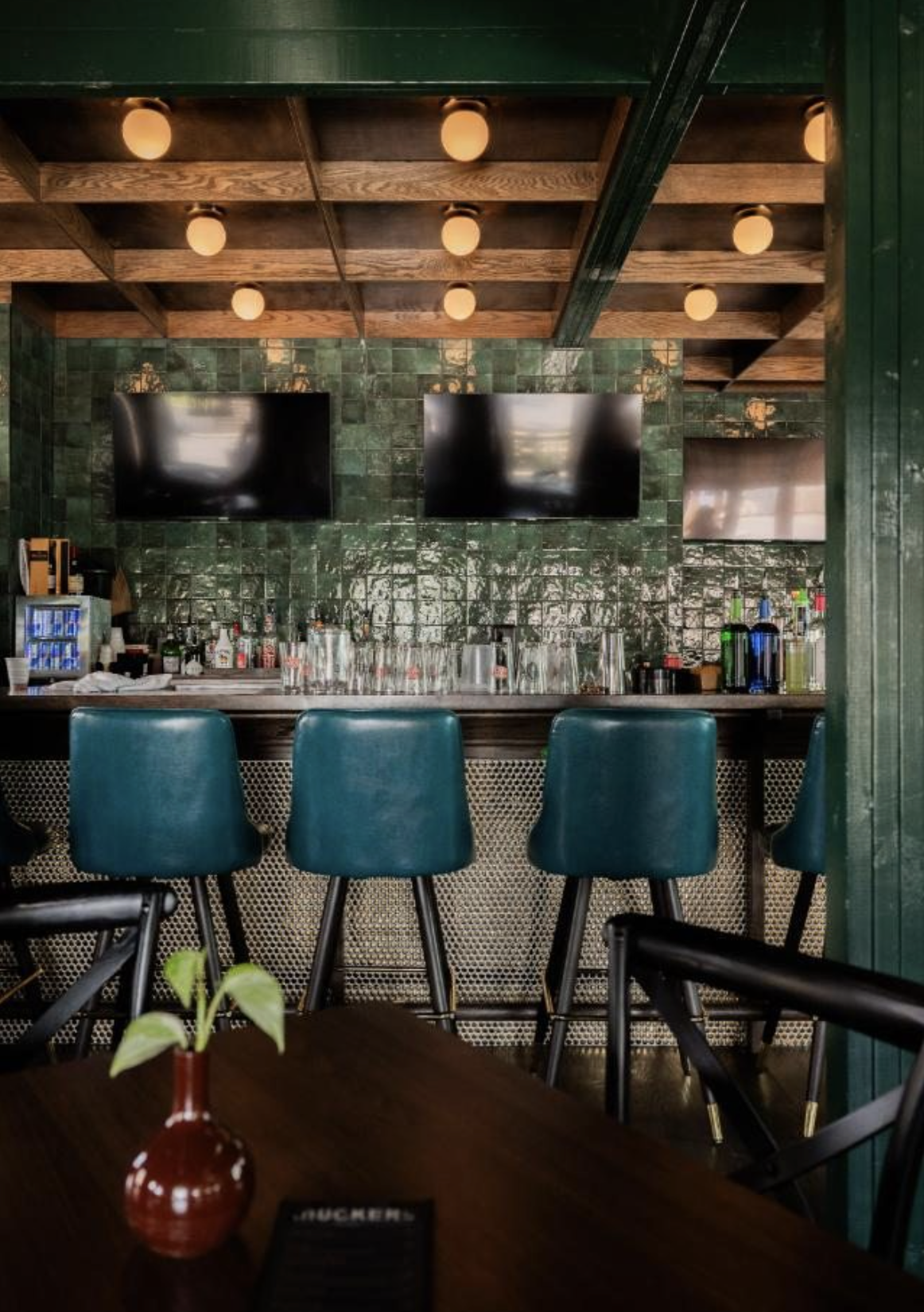 A cozy bar interior with a wooden ceiling and green tiled wall. Four teal bar stools are positioned in front of the bar, which has various bottles and glassware. Two large flat-screen TVs are mounted on the wall, and a small window is visible on the 