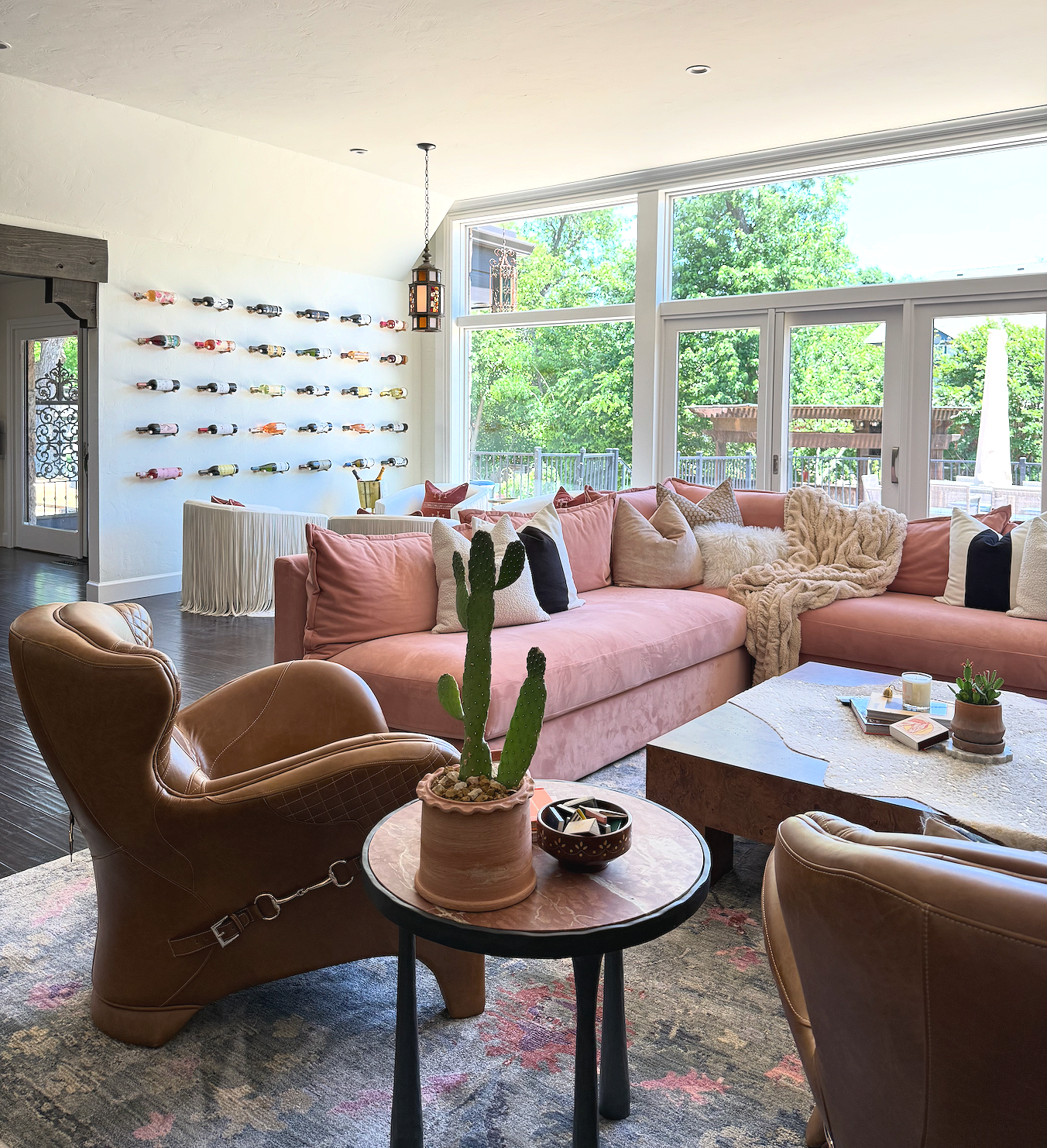 Living room with a pink sectional sofa, beige and black pillows, a furry blanket, large windows with a deck outside, a table with decorative items, a cactus plant, and a wall-mounted wine rack.
