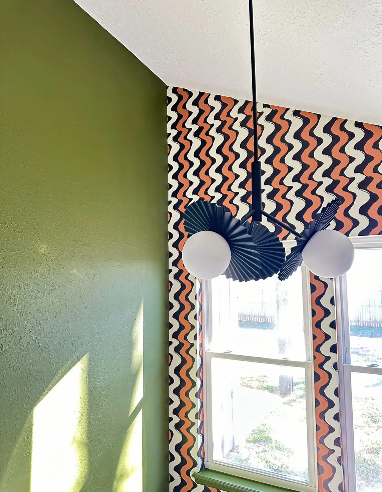 Decorative room featuring a green wall, a window with roller shade, and a string of paper lanterns or fans hanging from the ceiling. The wall beside the window has a multicolored zigzag pattern.