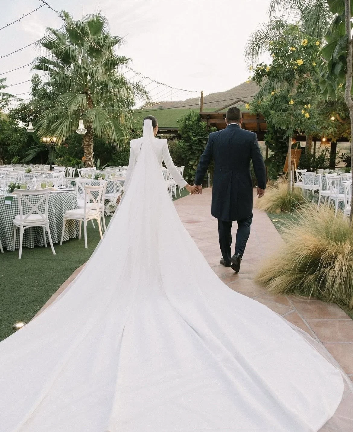 Una pareja de recién casados caminando de la mano en un lugar al aire libre decorado para una boda, con mesas vestidas y muchas plantas verdes.