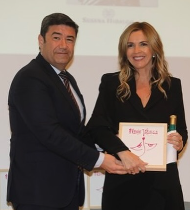 Una mujer y un hombre en un evento formal, la mujer sostiene un certificado con ilustraciones de senos y una botella de vino, ambos son de cabello rubio y vestimenta formal.