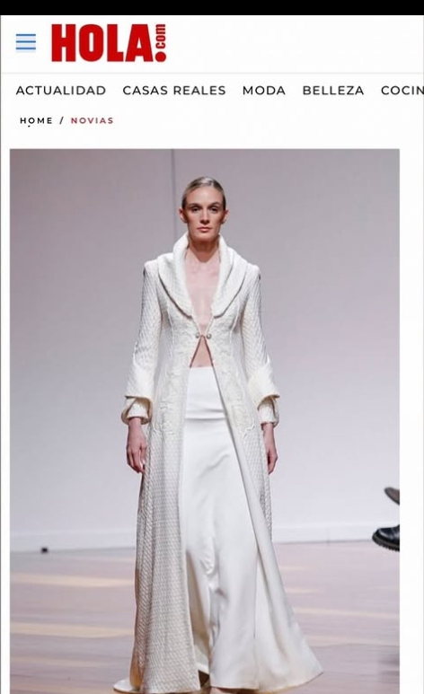 modelo en pasarela con vestido largo de color blanco y abrigo de colores claros, fondo gris y logo de 'Hola.com' en la parte superior.