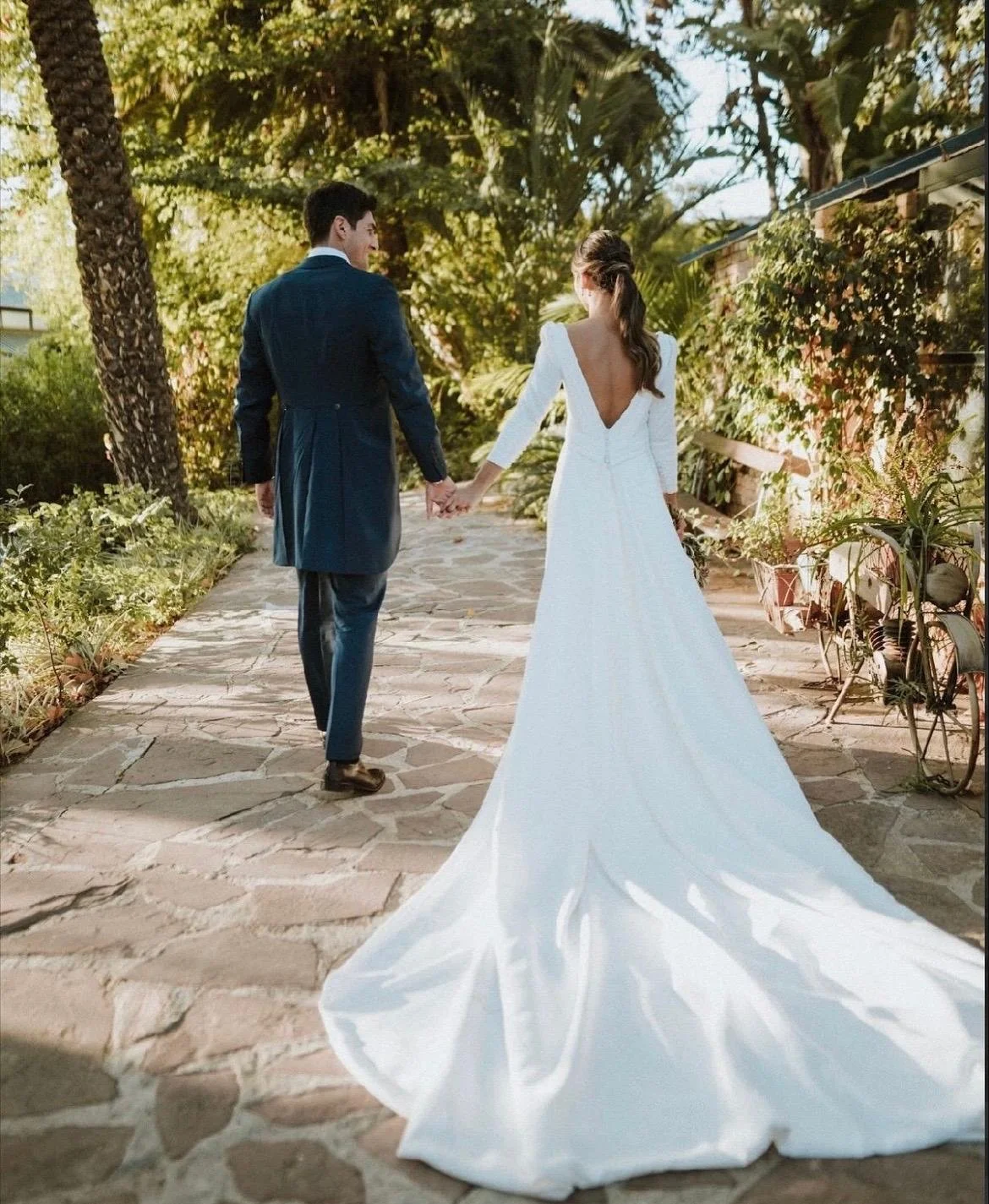 Pareja de recién casados caminando de la mano en un sendero exterior rodeado de vegetación, con ella llevando un vestido de boda blanco de larga cola y él un traje oscuro.